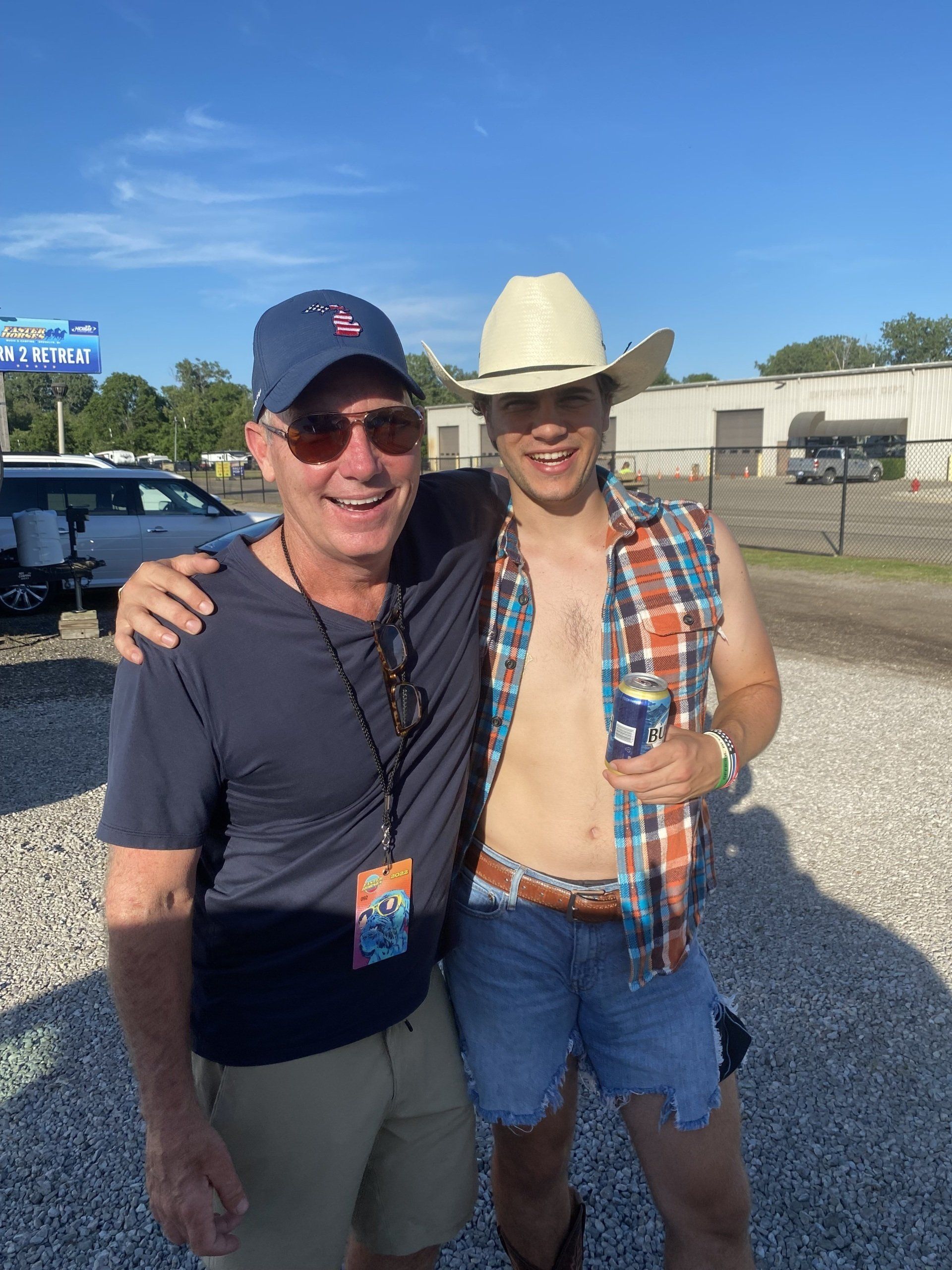 Two men are posing for a picture in a gravel lot . one of the men is wearing a cowboy hat.