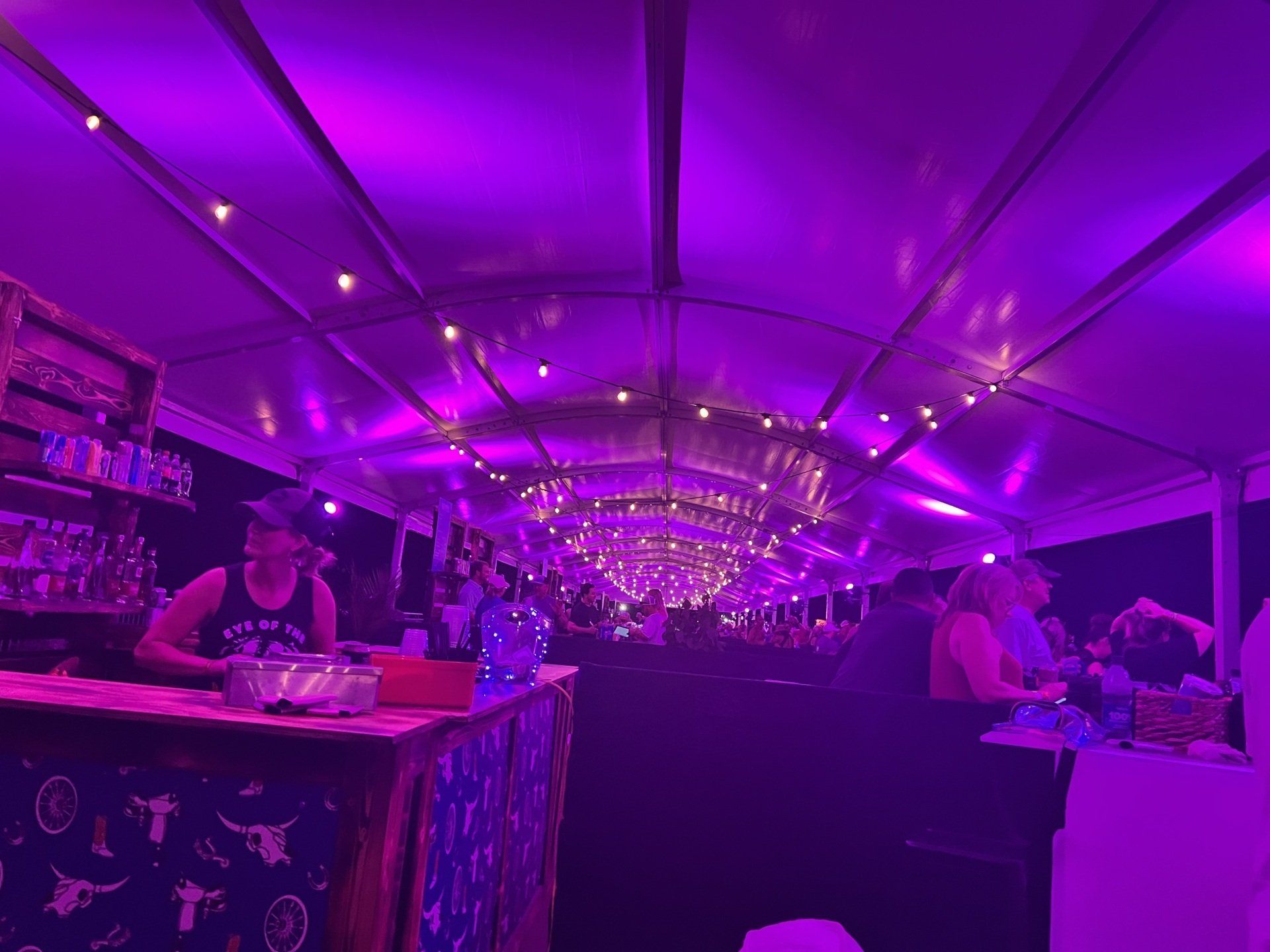A group of people are sitting at a bar under a tent with purple lights.