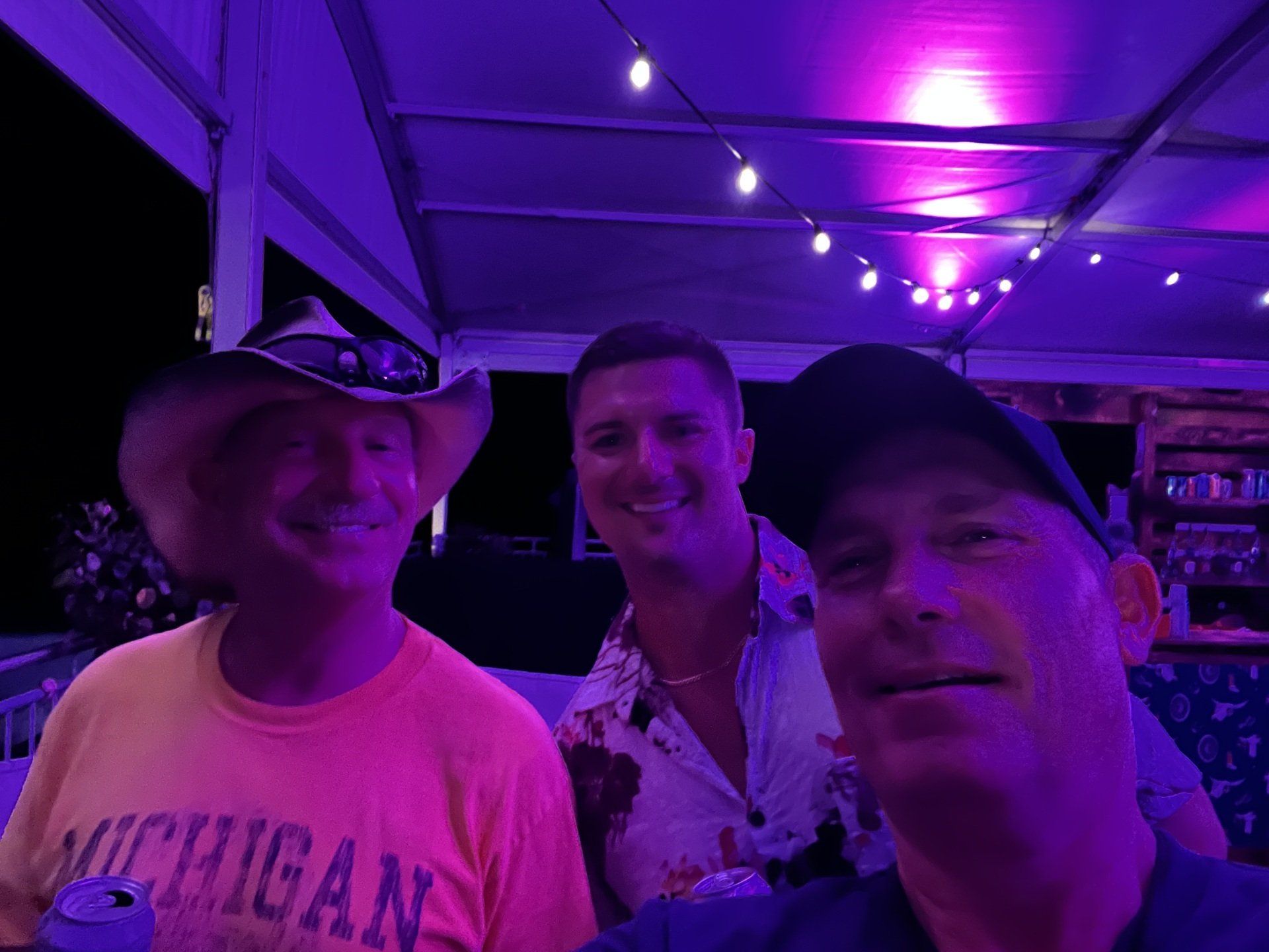 Three men are posing for a picture in front of a purple light . one of the men is wearing a michigan shirt.