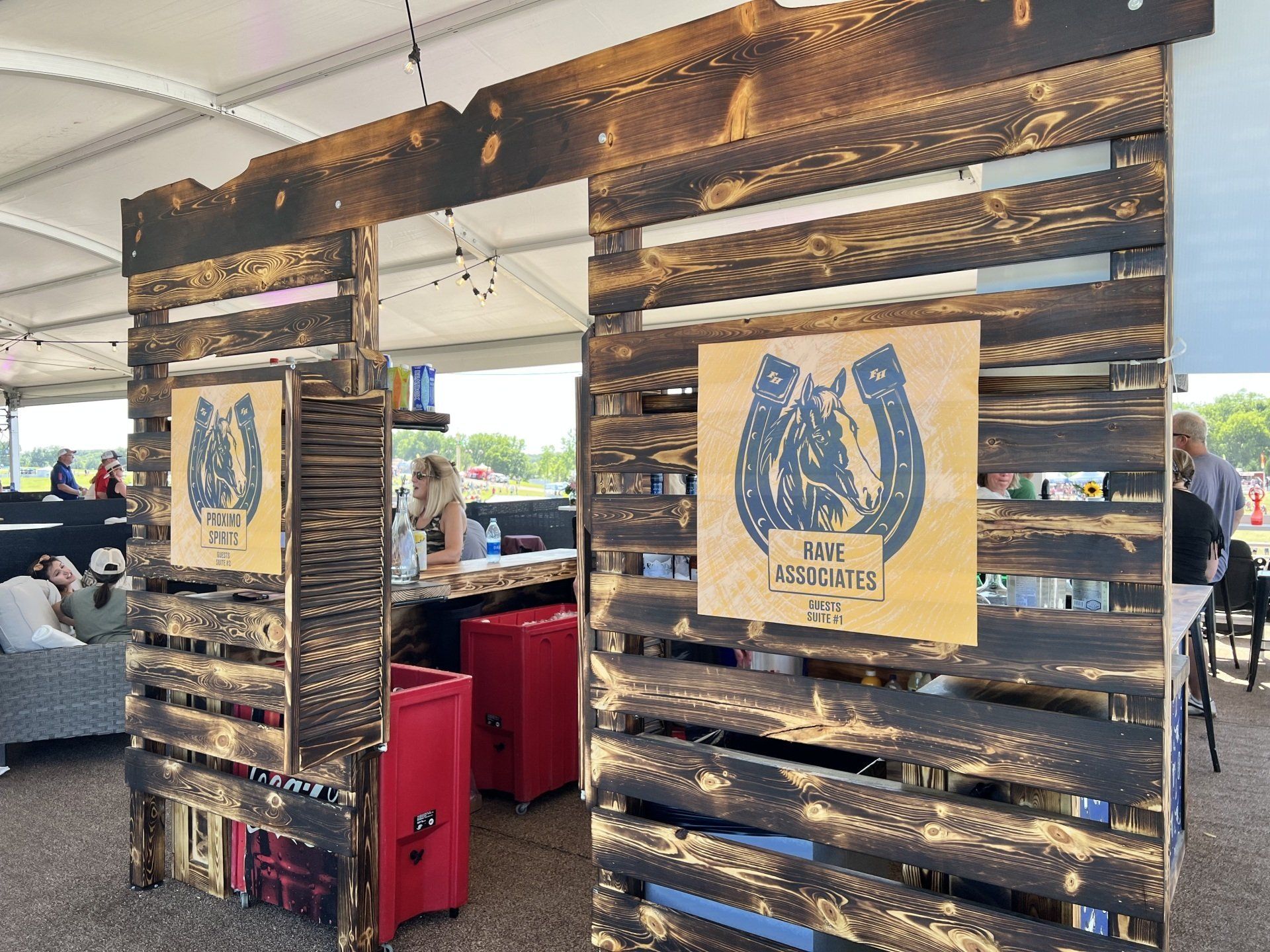 A group of people are sitting at tables under a tent behind wooden pallets.