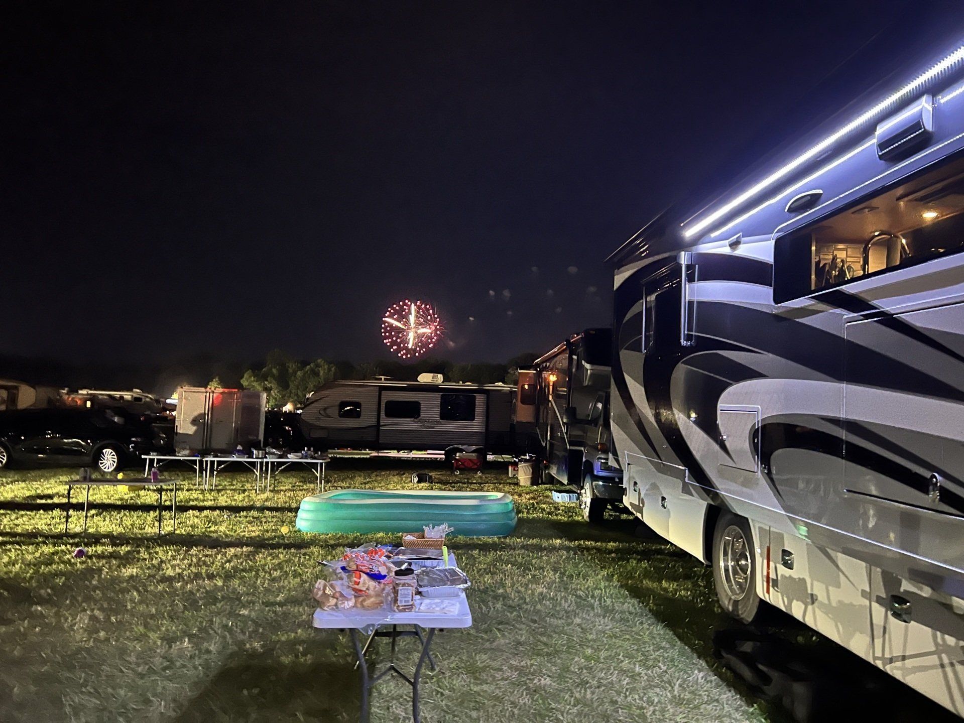 A rv is parked in a field at night with fireworks in the background.