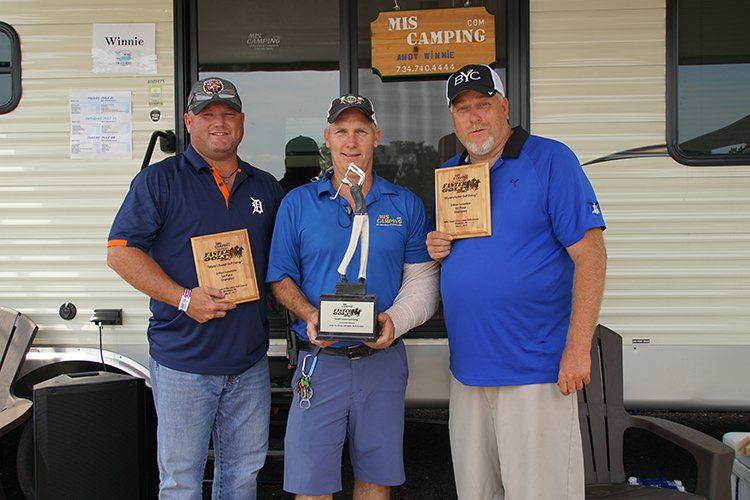 Three men are standing in front of a trailer holding trophies.
