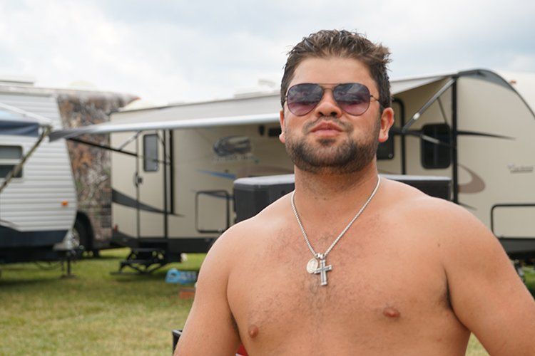 A shirtless man wearing sunglasses and a cross necklace is standing in front of a trailer.