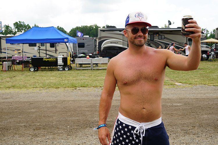 A shirtless man is standing in a field holding a can of beer.