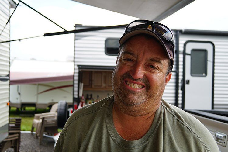 A man wearing a hat and sunglasses is smiling in front of a trailer.