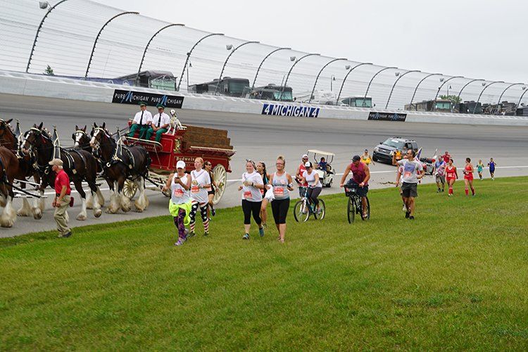 A group of people are running in front of a horse drawn carriage on a race track.