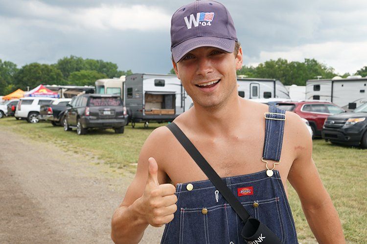 A shirtless man wearing overalls and a hat is giving a thumbs up