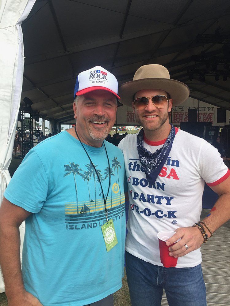 Two men are posing for a picture in front of a tent . one of the men is wearing a born party rock shirt.