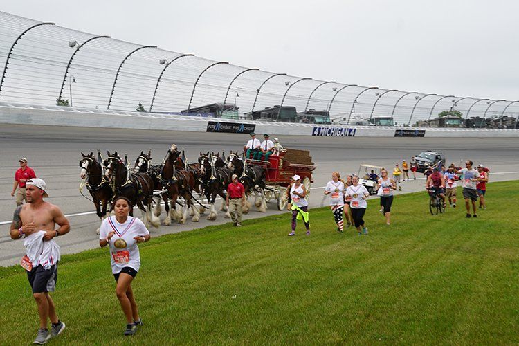 A group of people are running in front of a horse drawn carriage on a race track.