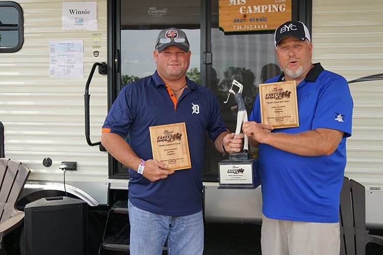 Two men are holding trophies in front of a trailer.