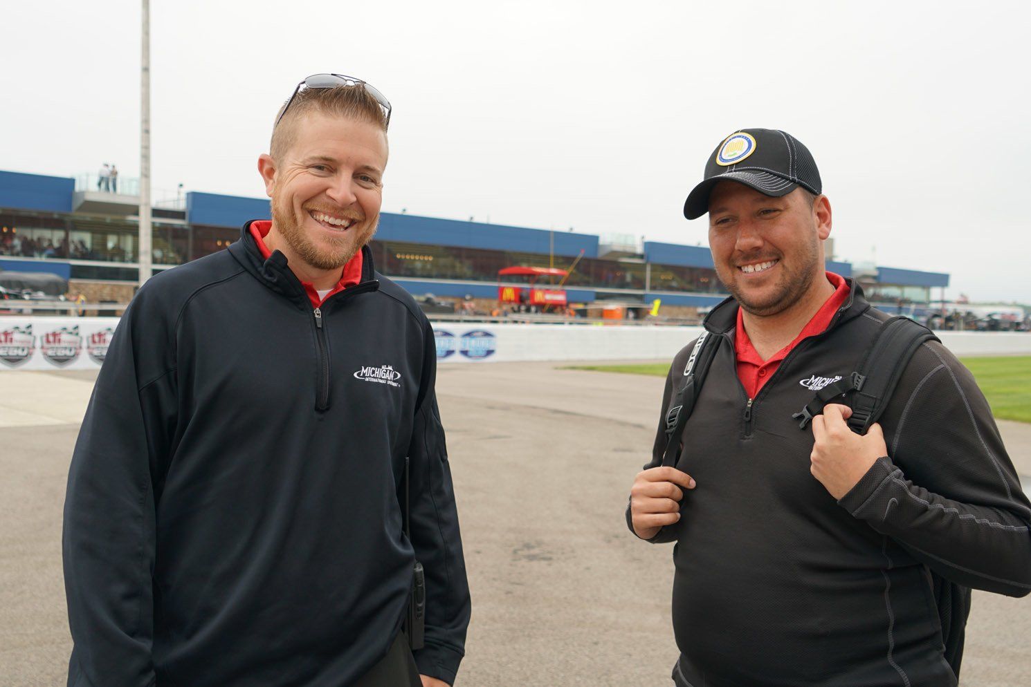 Two men are standing next to each other in front of a race track.