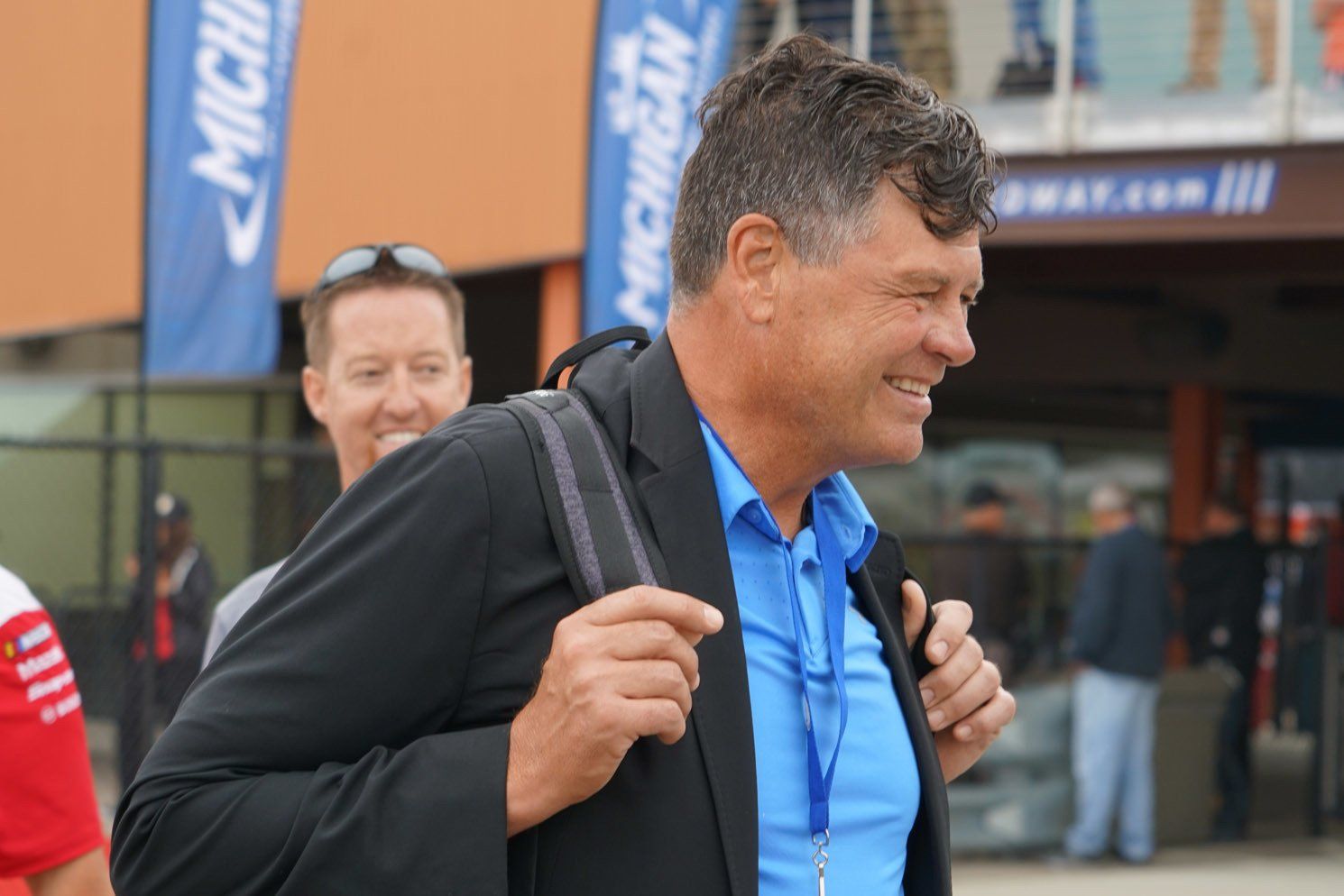 A man in a blue shirt and black jacket is smiling while carrying a backpack.