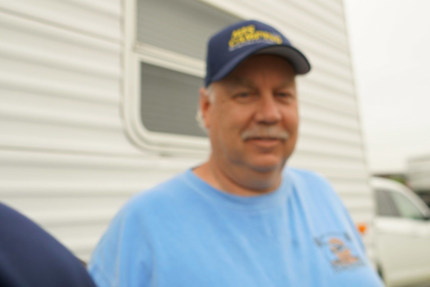 A man wearing a blue shirt and a blue hat stands in front of a trailer