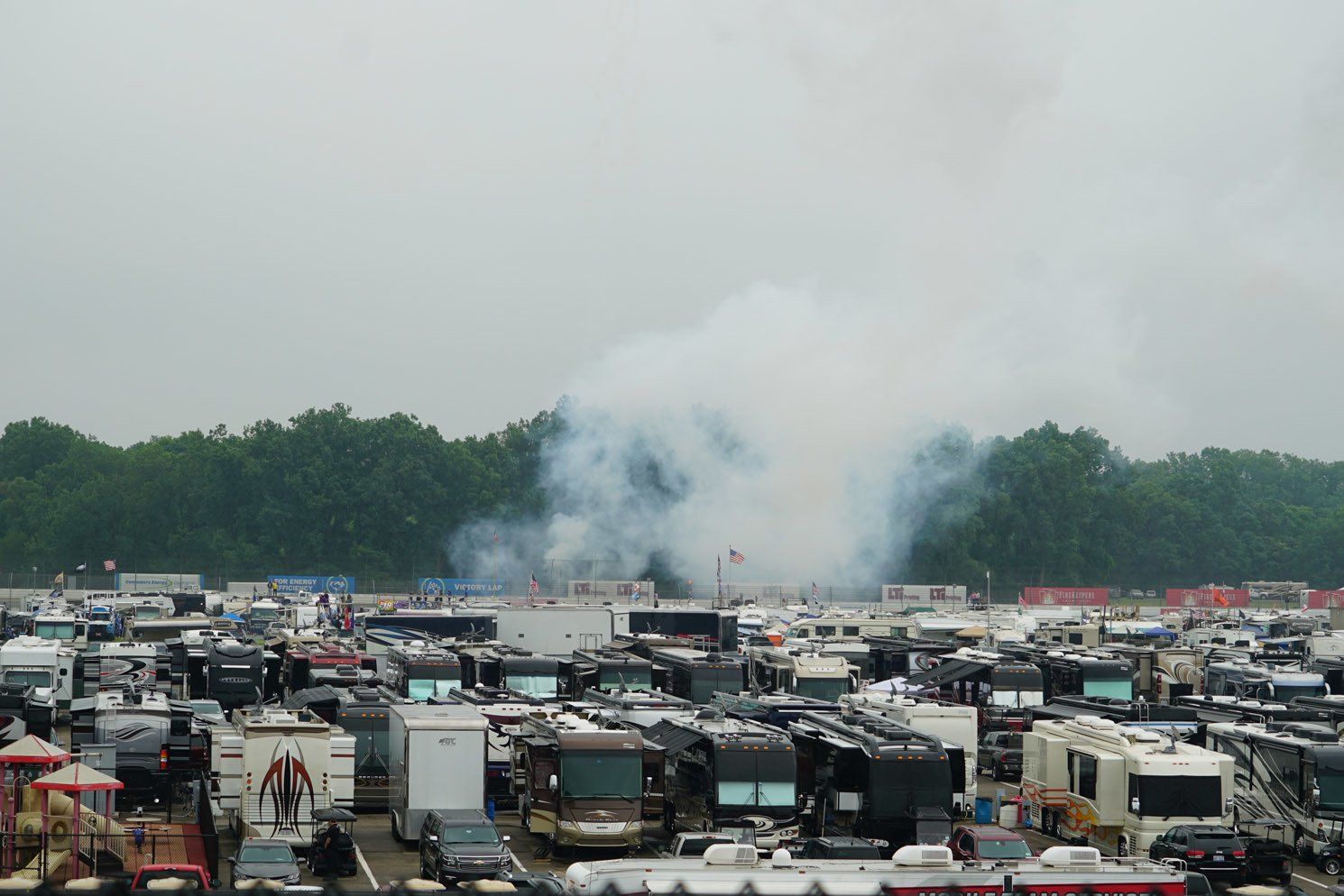 A lot of vehicles are parked in a lot with smoke coming out of the trees