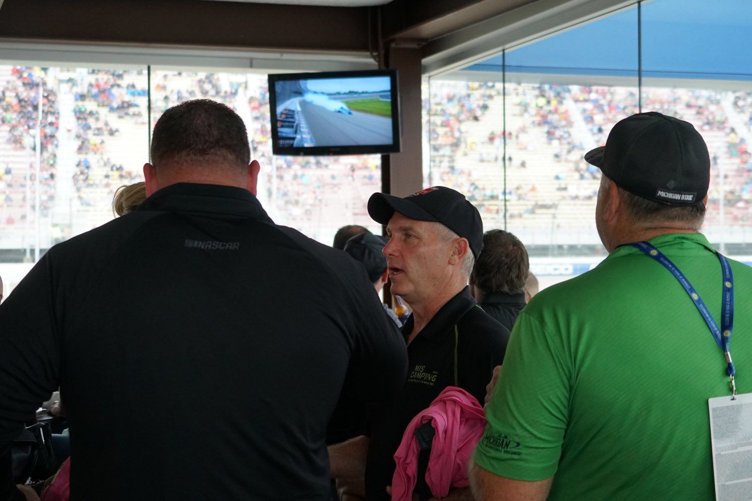 A group of men are standing in a room watching a race on a television.