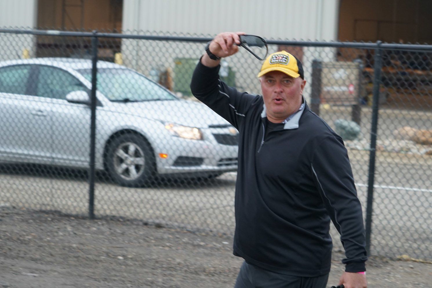 A man wearing a black shirt and a yellow hat is standing in front of a chain link fence with a car in the background