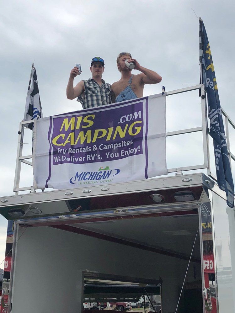 Two men are standing on top of a trailer with a mis camping sign.