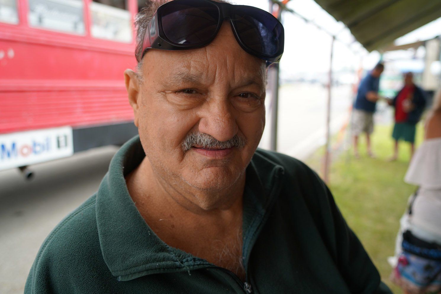 A man wearing sunglasses is smiling in front of a red mobil bus
