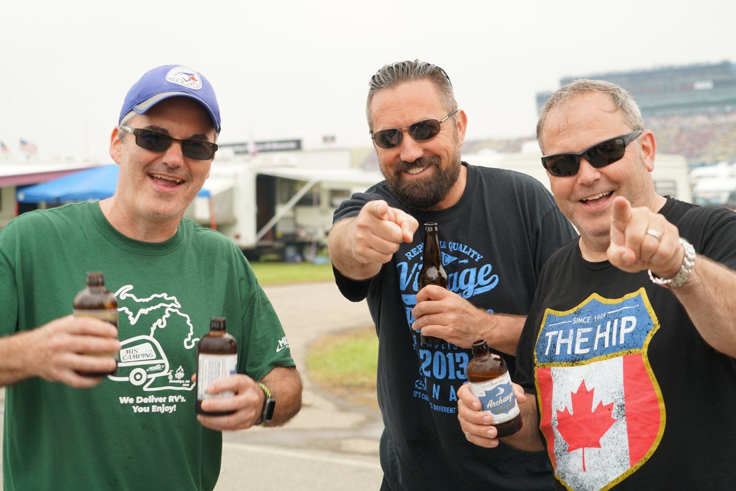 Three men are standing next to each other holding bottles of beer and pointing at the camera.