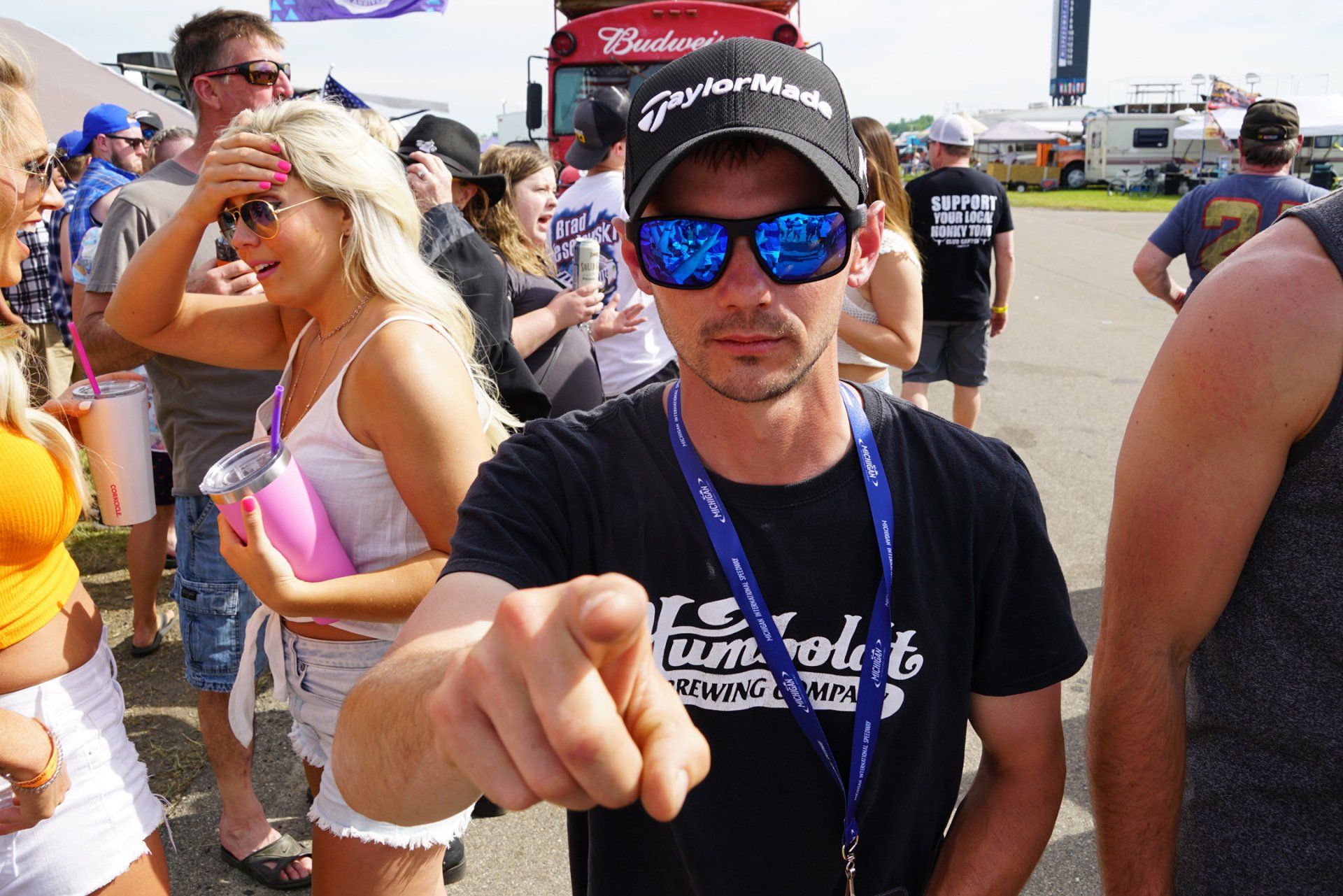 A man wearing sunglasses and a budweiser hat points at the camera