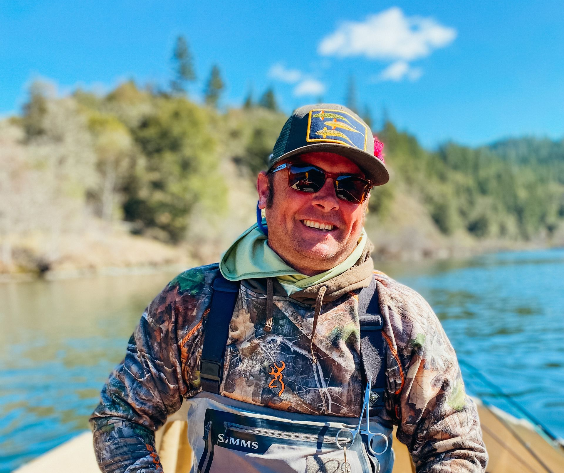 Man in fishing gear smiling, rowing a boat on a lake; sunny day, trees in background.