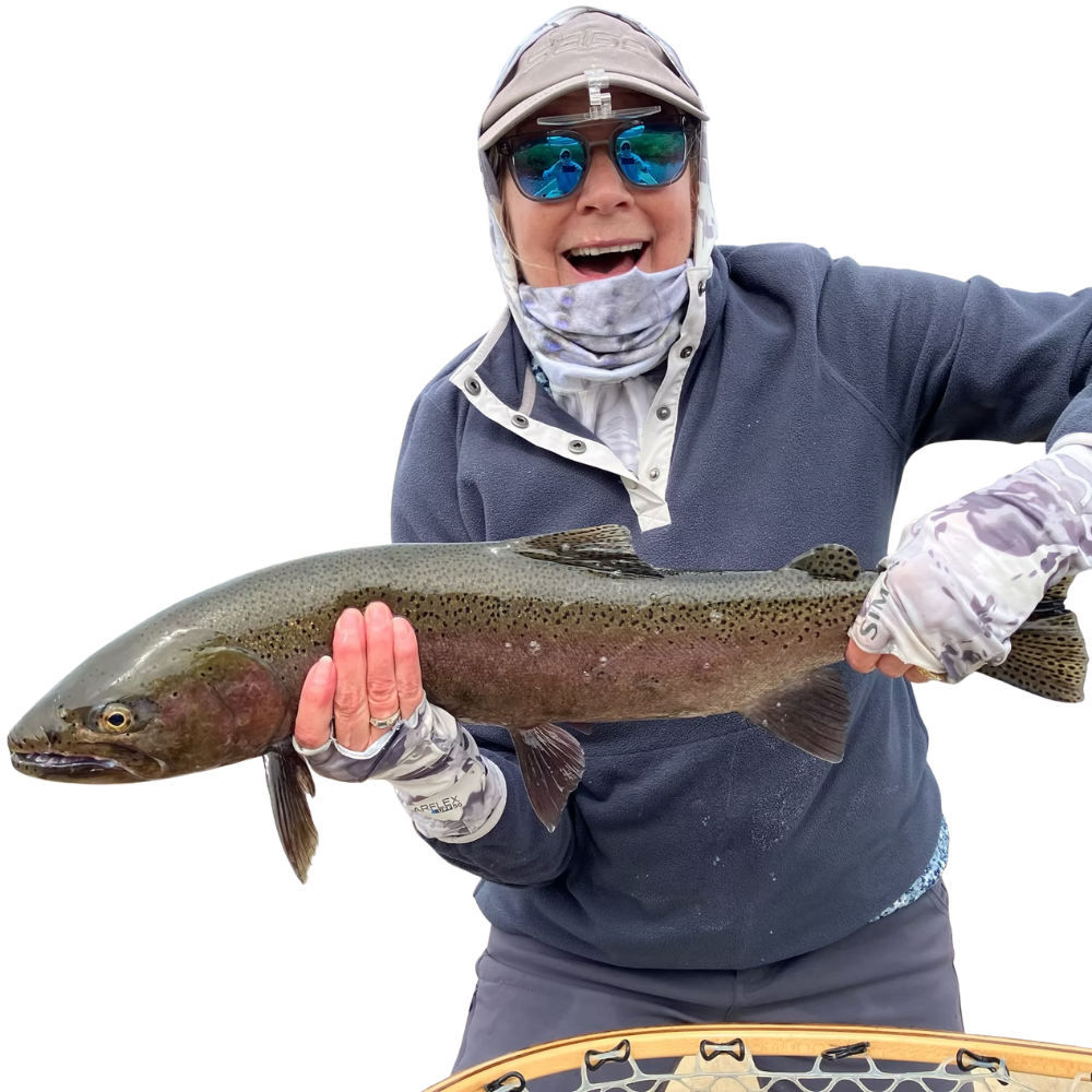 Woman wearing fishing gear holds a large rainbow trout, smiling. Outdoor setting, grey and blue tones.