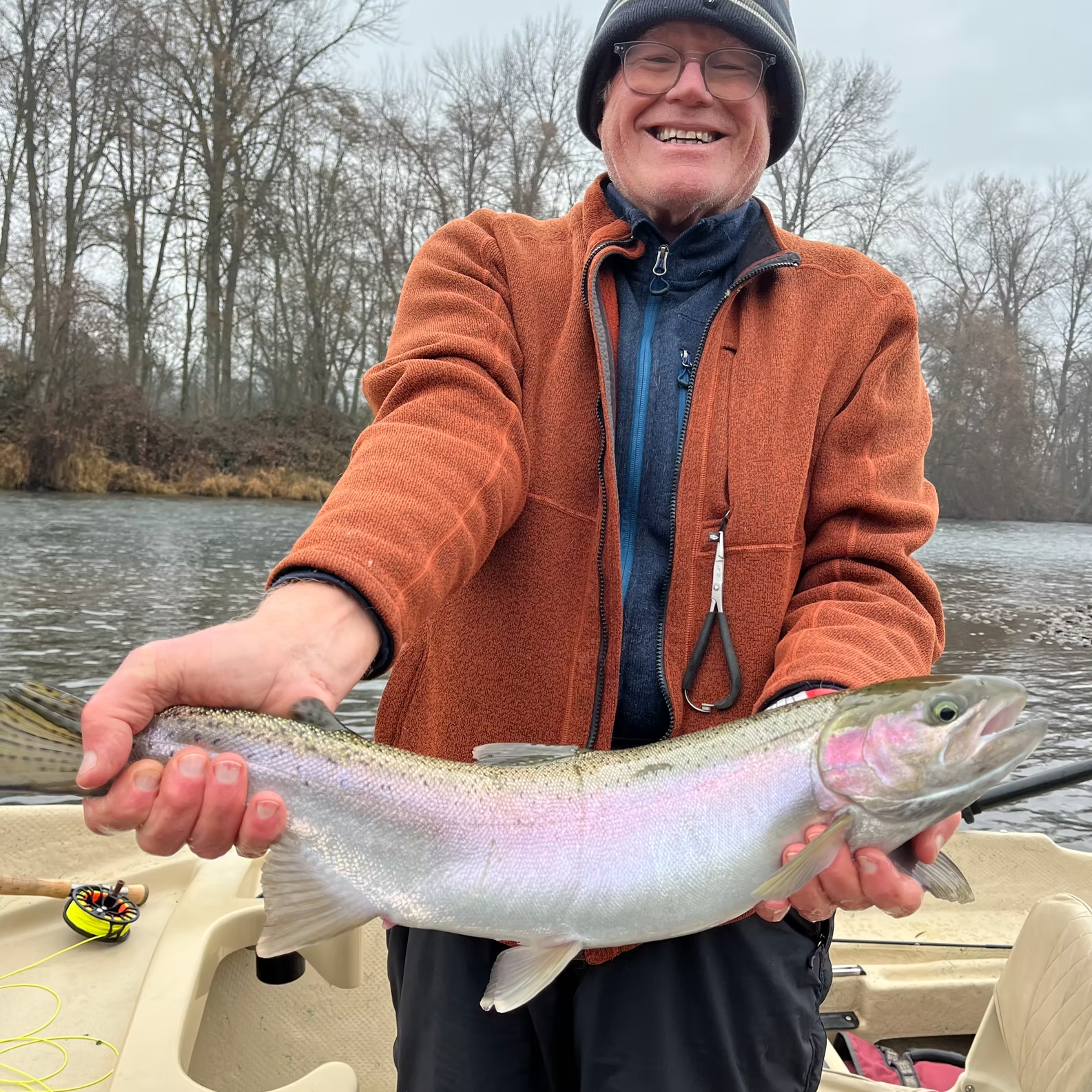 Smiling man holding a rainbow trout in a boat, near a river with trees.