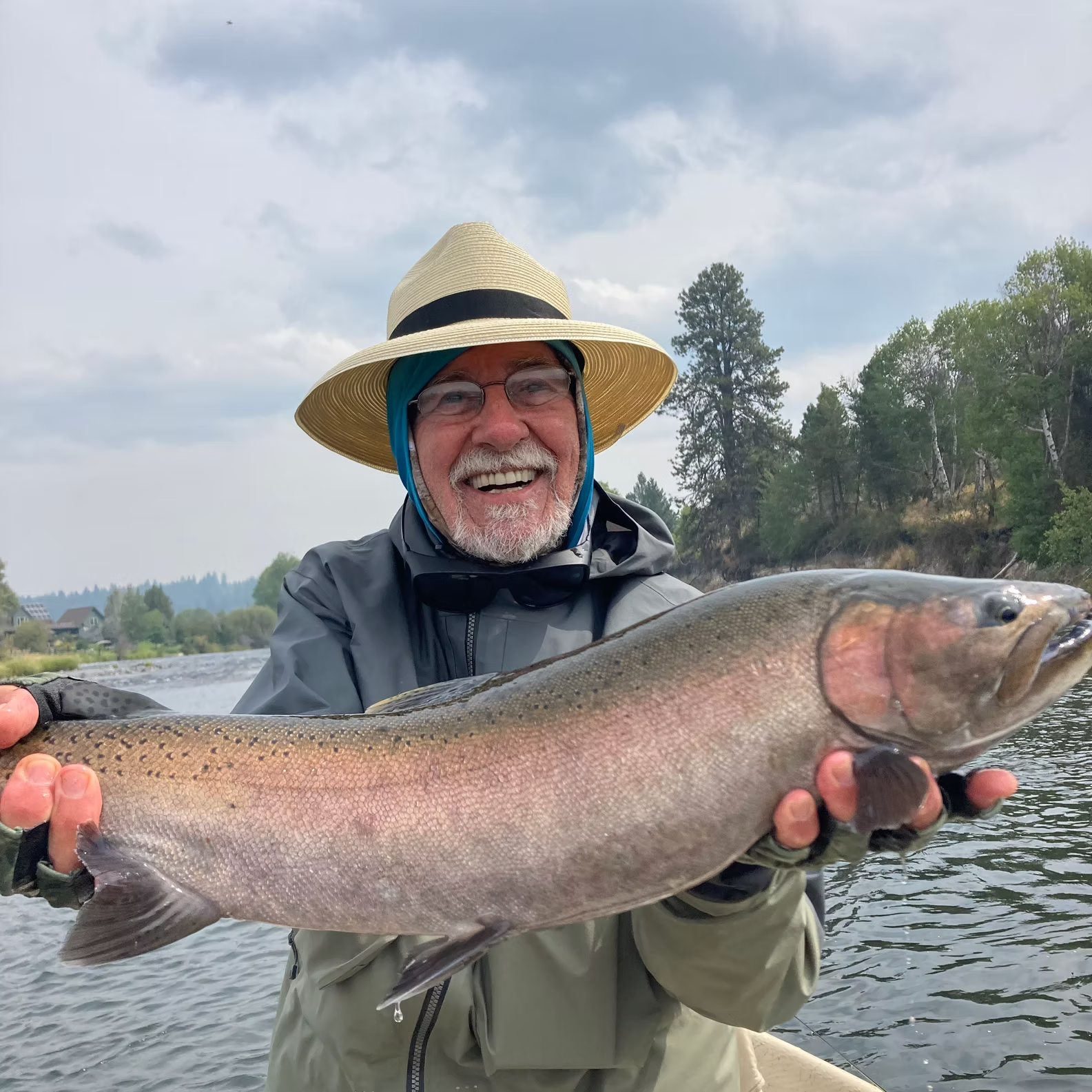 Man in hat holds up large rainbow trout next to river, smiling.