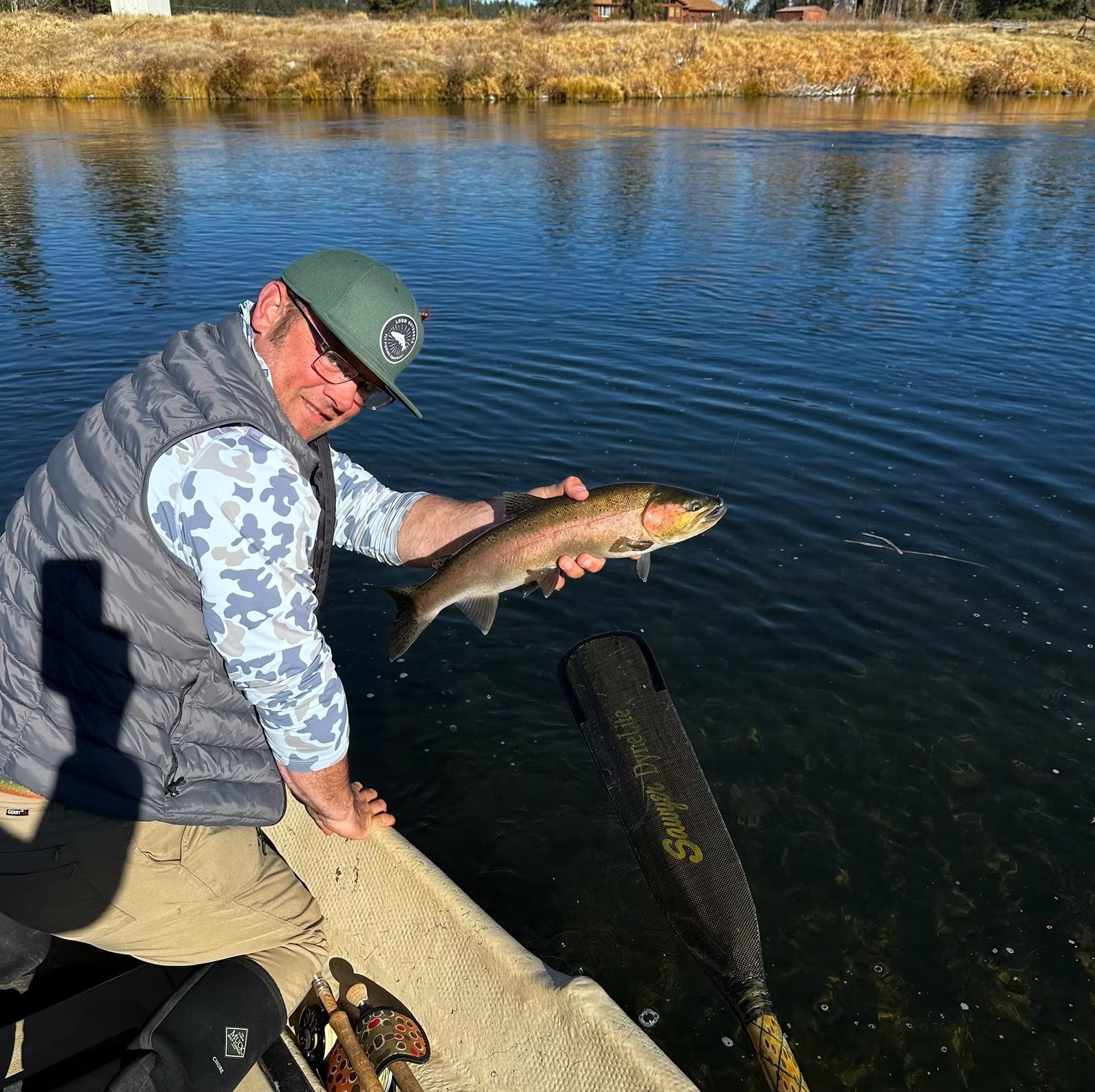 Man holding a fish in a boat on a river, sunny day. He wears a hat and vest.