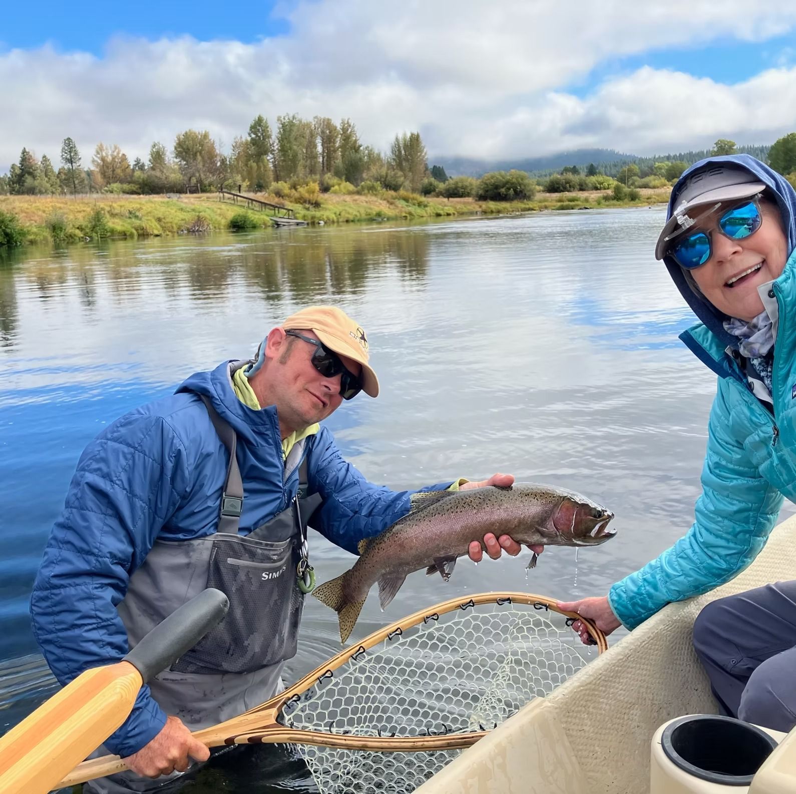 Two people on a boat, holding a large trout. One has a net, blue water and sky, green shoreline.