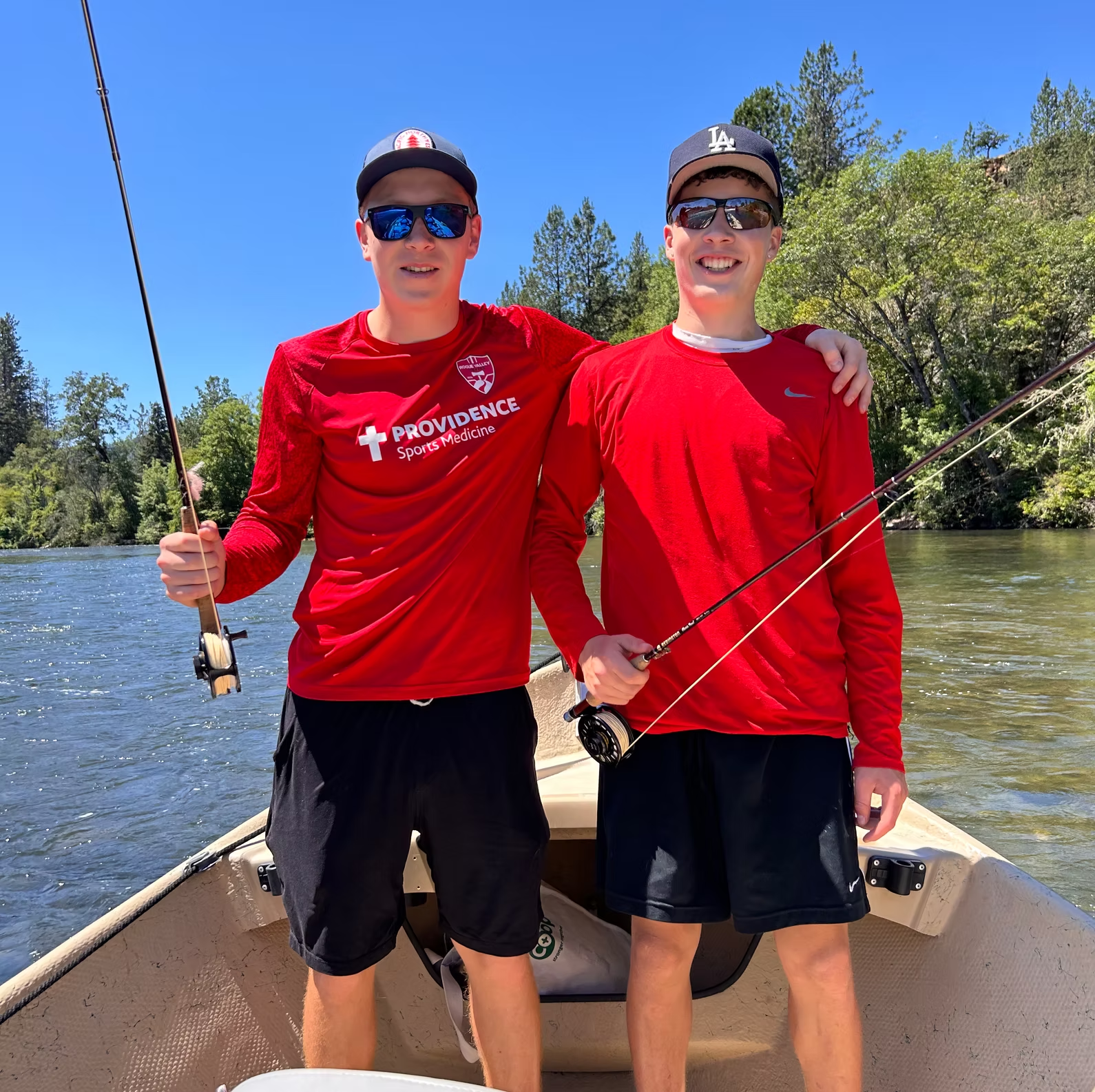 Two young men in a boat, fly fishing on a river. Sunny day, wearing red shirts and sunglasses.