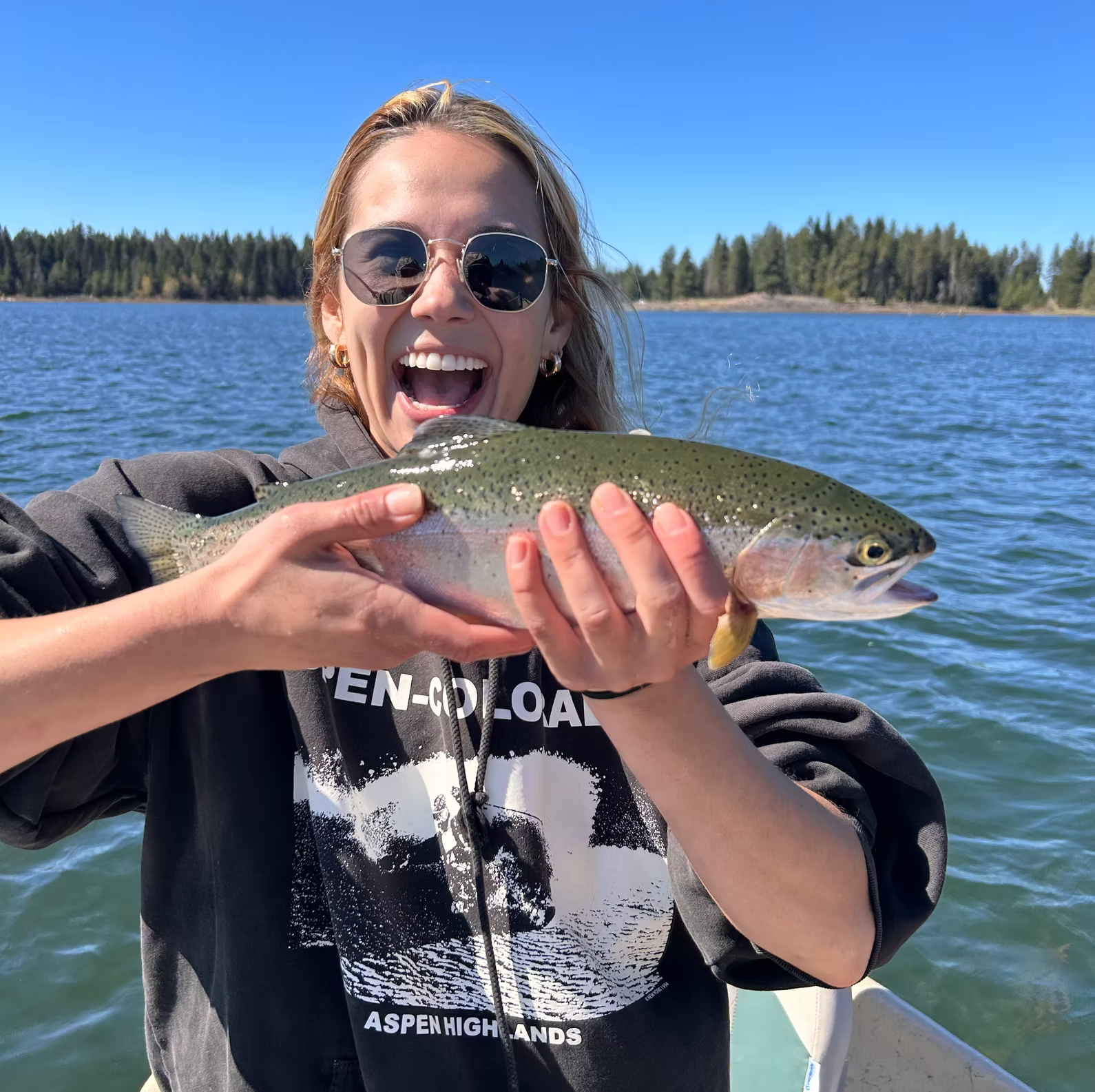 Woman in sunglasses smiles, holding rainbow trout. Blue lake and green trees in the background.