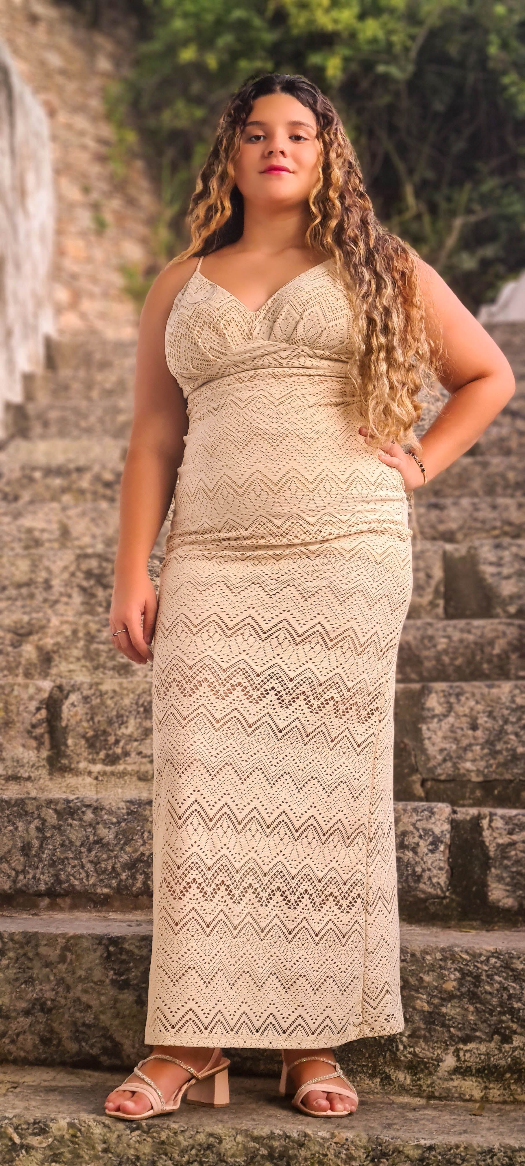Mulher com um vestido creme está em degraus de pedra, com a mão no quadril, olhando para a câmera.