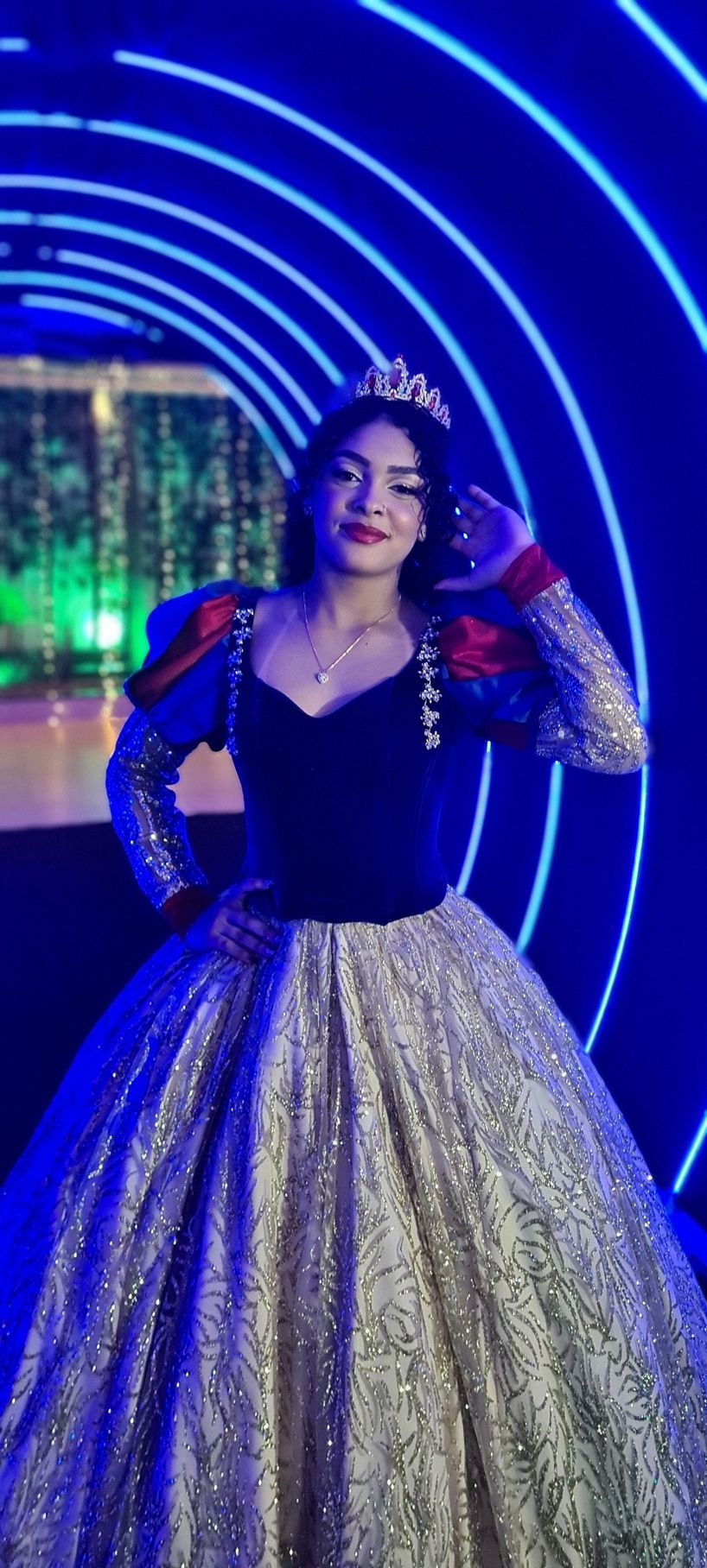 Mulher com vestido inspirado em Branca de Neve posa em um túnel iluminado em azul. Ela usa uma coroa e sorri, com a mão no ombro.