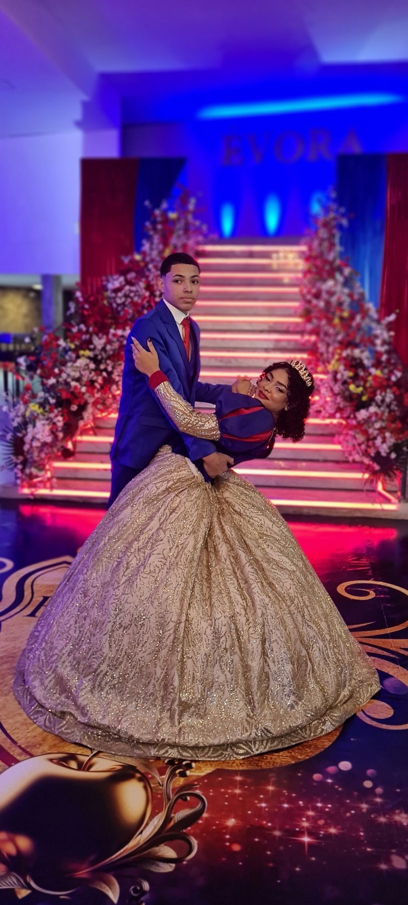 Casal dançando em um cenário glamouroso; mulher de vestido dourado se inclina para trás enquanto homem de terno azul a abraça.