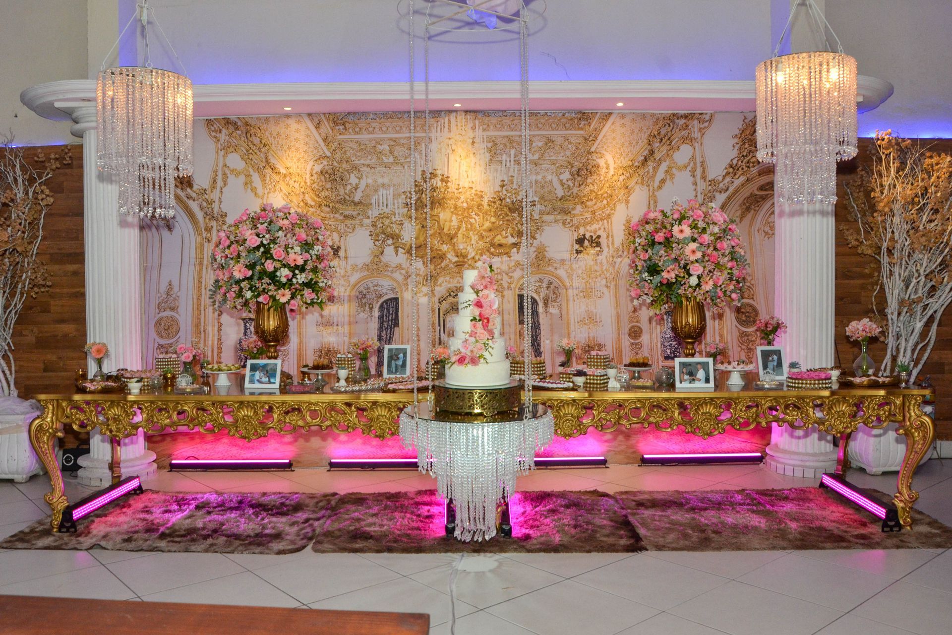 Mesa de sobremesas elegante com bolo, flores e decoração dourada, iluminada com luzes rosa e roxas.
