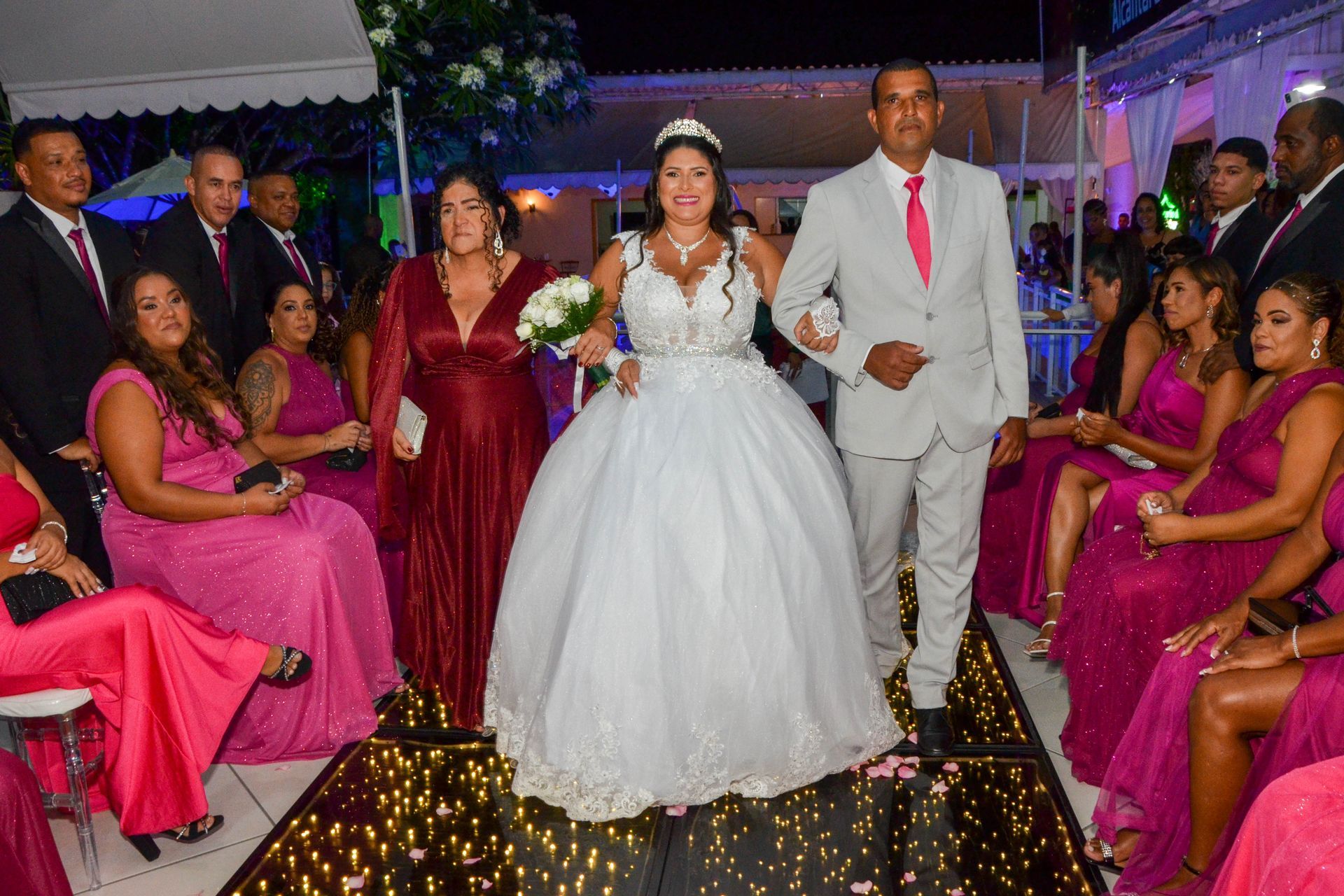 Noiva caminhando pelo corredor com um dos pais, cercada pelos convidados em um espaço decorado.