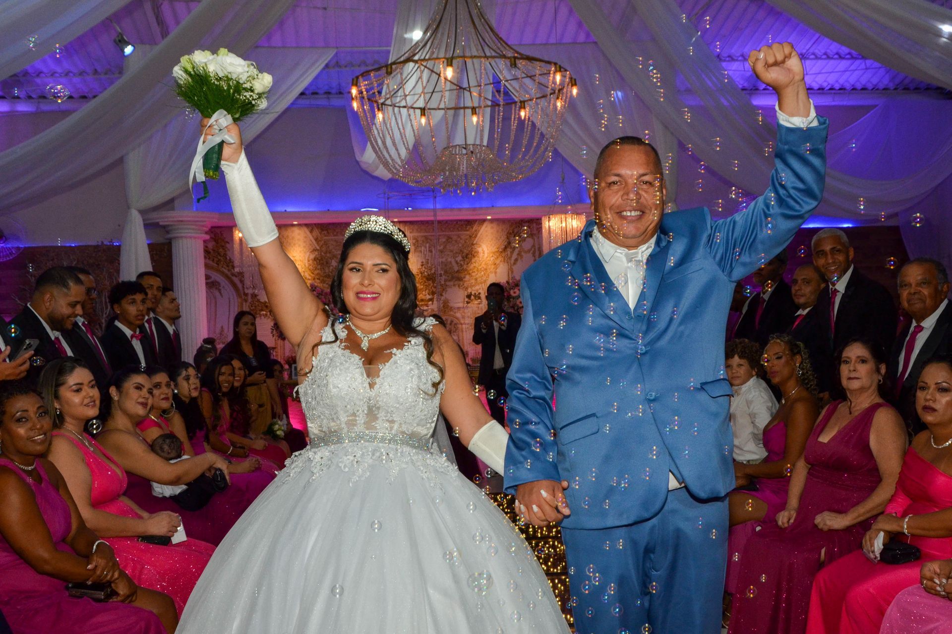 Recém-casados levantam os braços, comemorando entre os convidados em uma recepção de casamento.