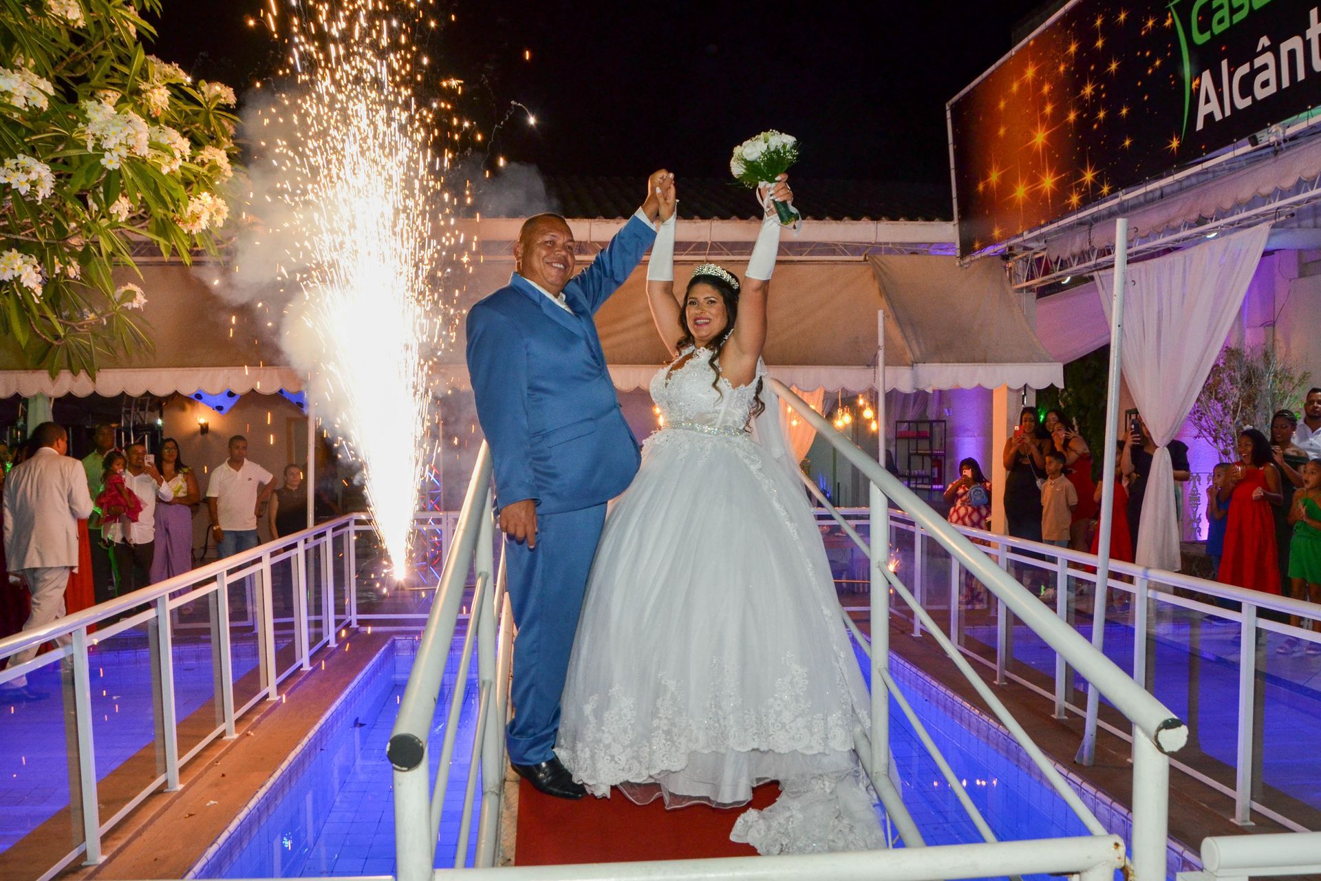 Recém-casados na ponte com fogos de artifício; mulher de vestido branco, homem de terno azul, noite.
