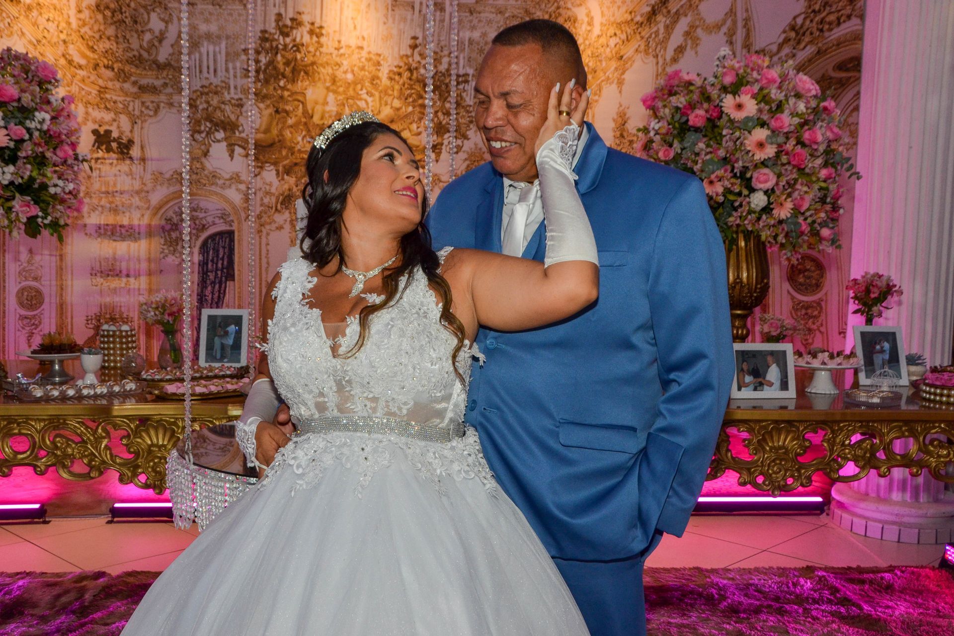 Noivos se abraçam em um casamento. Ela usa um vestido branco e tiara; ele, um terno azul. Iluminação rosa e decoração floral.