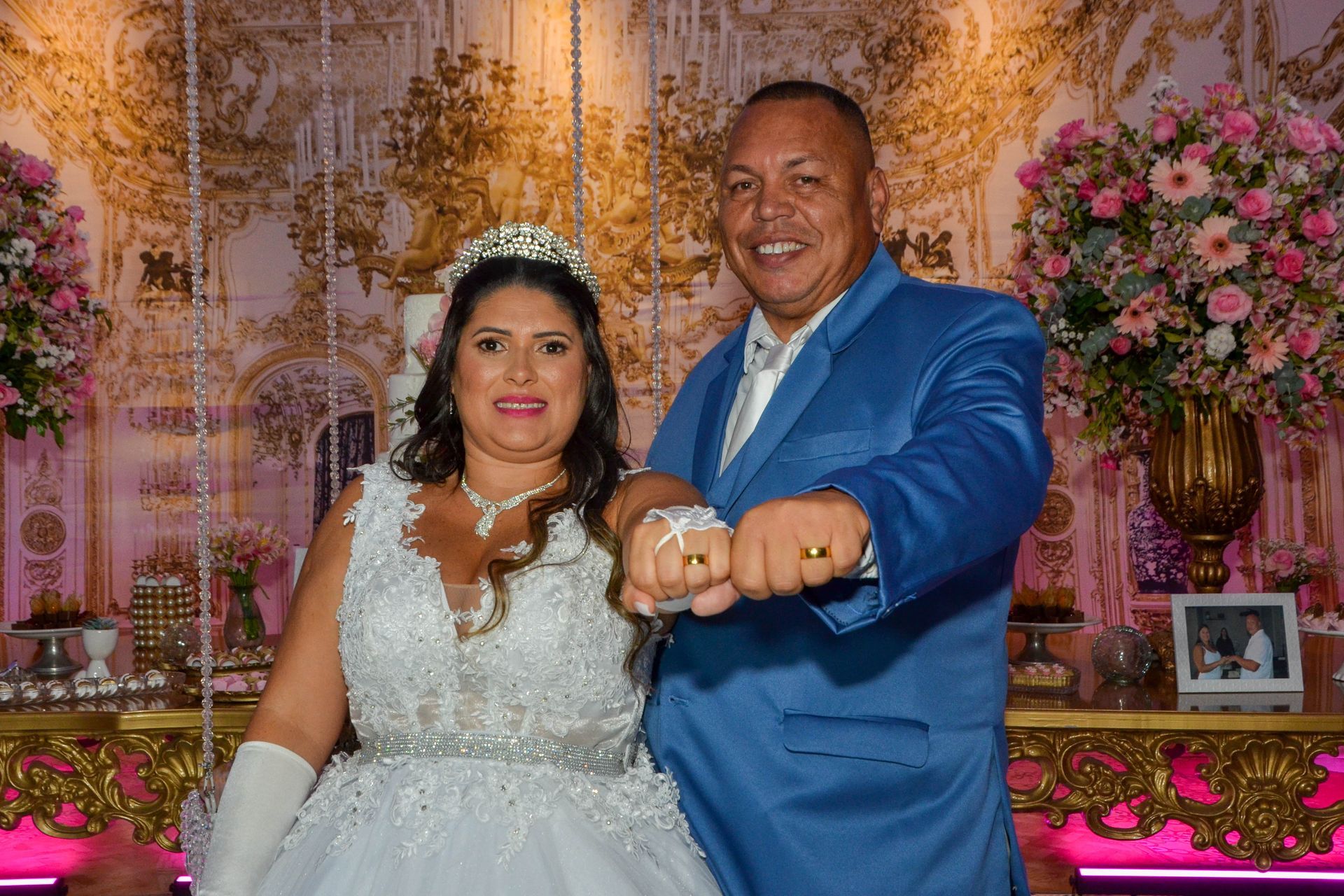 Noivos sorrindo, mostrando alianças. Quarto decorado, flores, bolo, detalhes dourados.