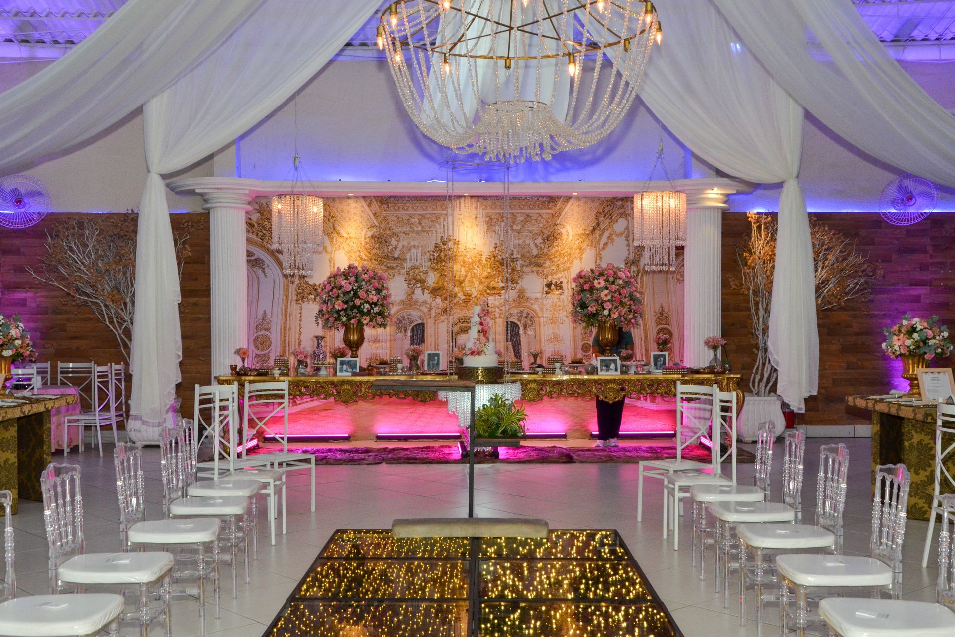 Espaço elegante para eventos com teto drapeado, mesa decorada e cadeiras transparentes.