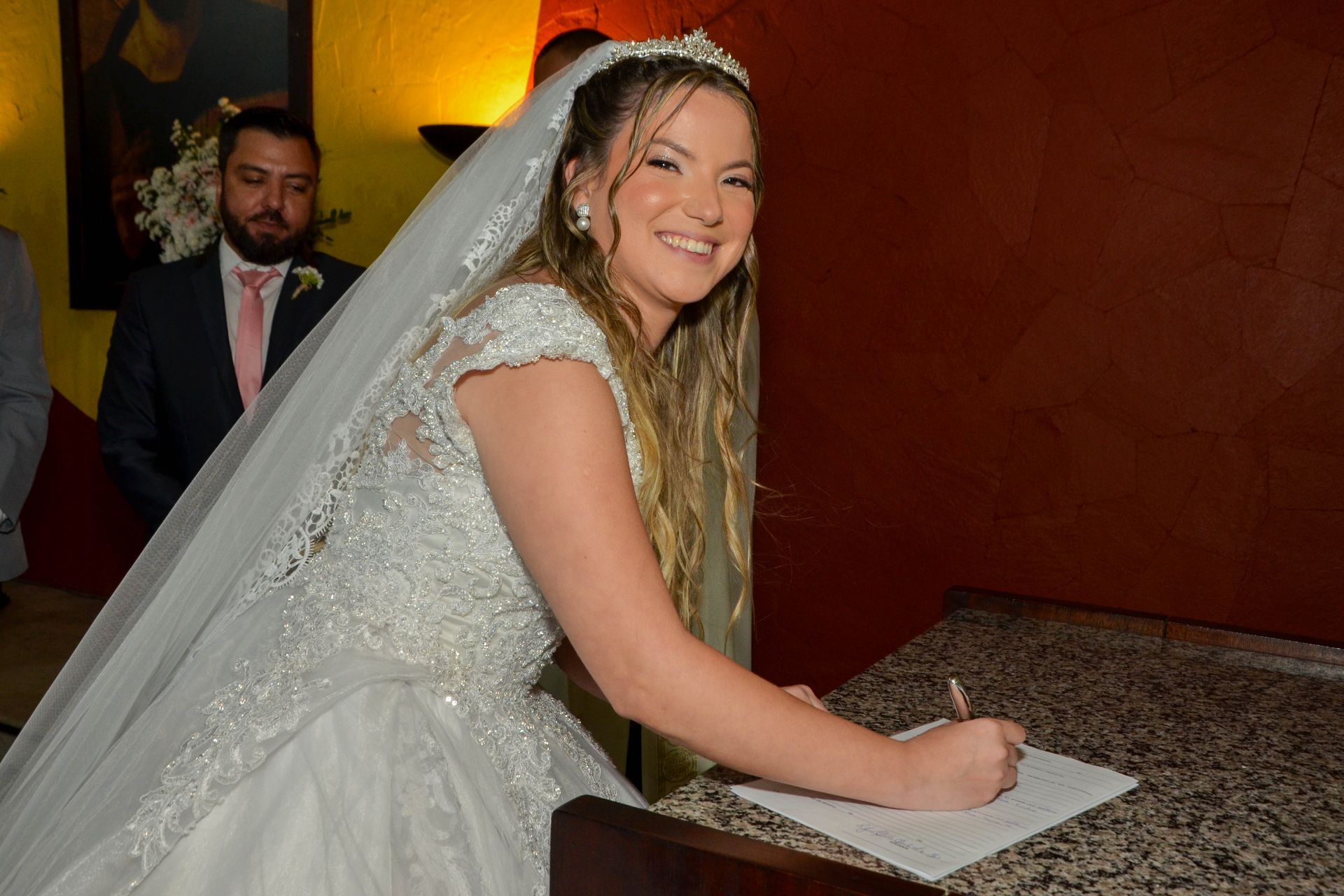 Noiva vestida de noiva assinando um documento à mesa, sorrindo. Noivo de terno observa ao fundo.