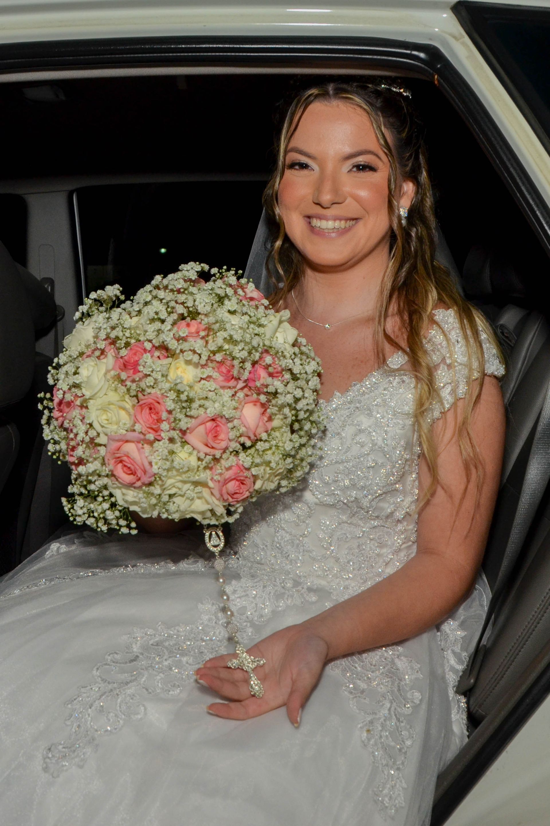 Noiva em vestido de noiva branco com contas sorrindo, segurando um buquê, saindo do carro.