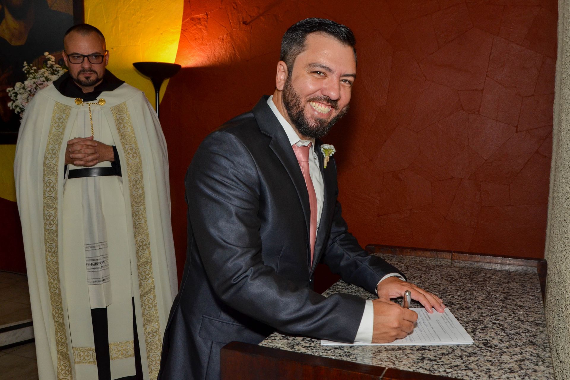 Homem de terno assina documento, sorrindo; padre de paramentos observa.