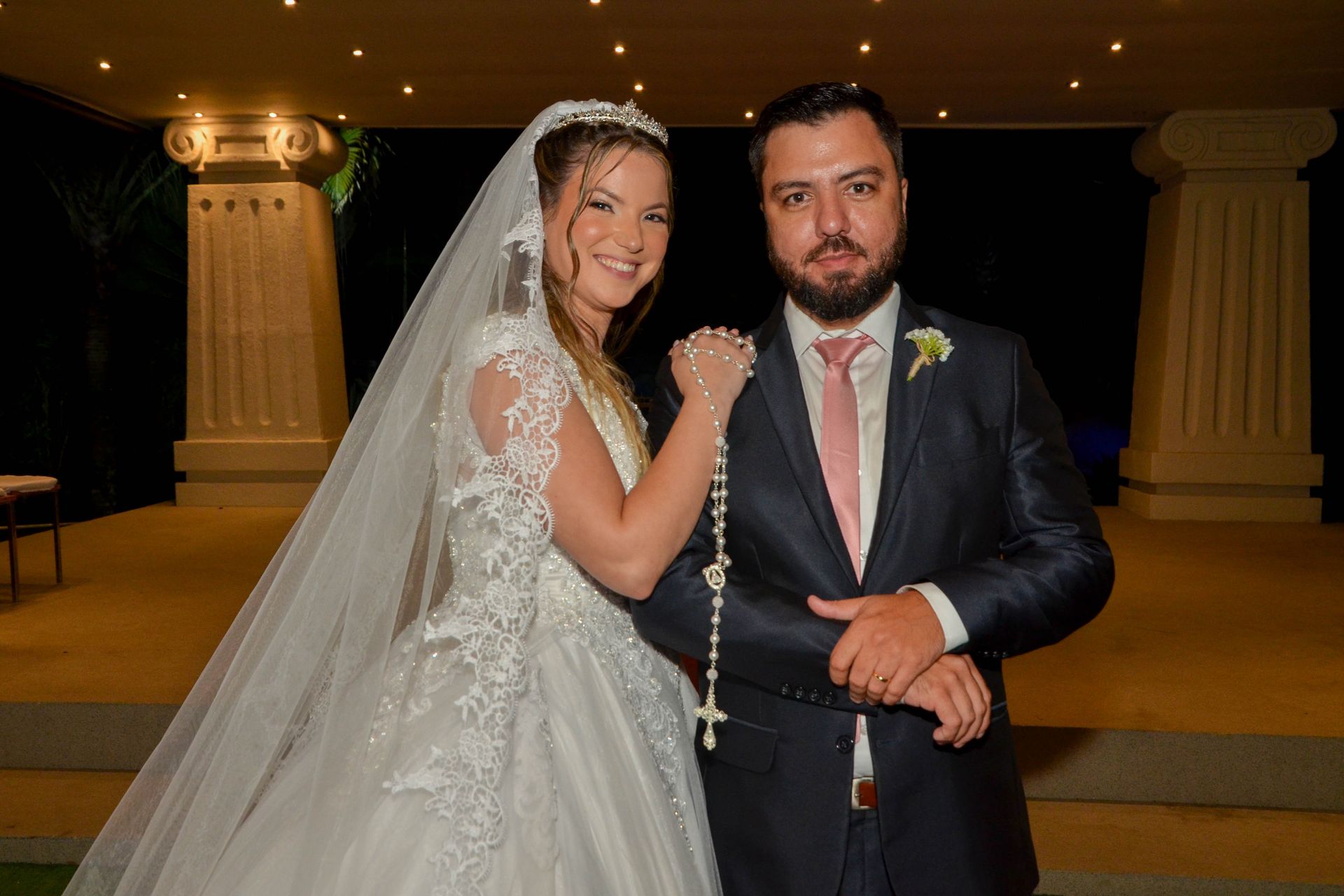 Noivos sorrindo para a câmera, cerimônia de casamento, vestido branco, terno escuro, pérolas.