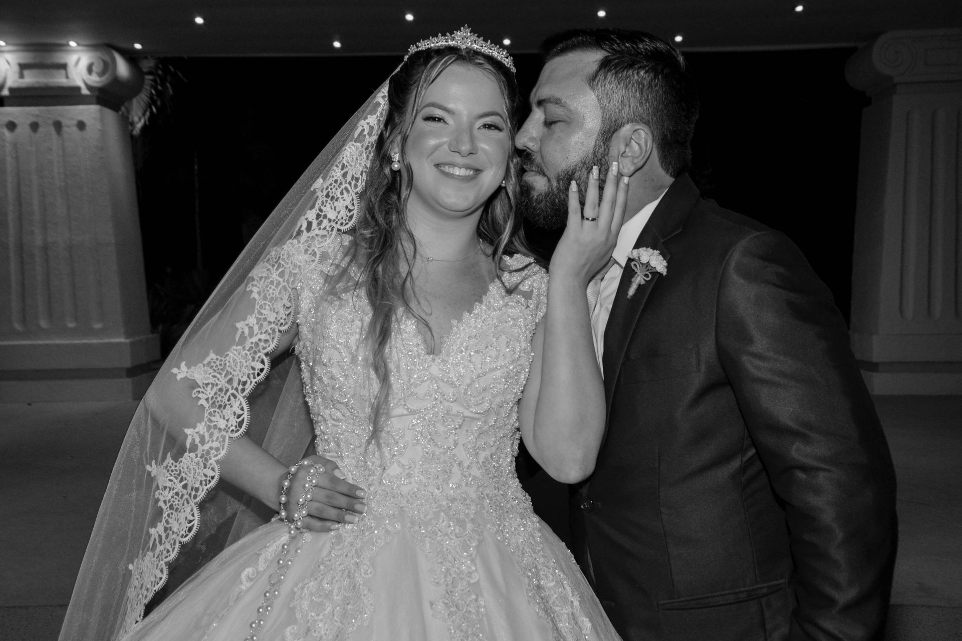 Noivos sorrindo, em trajes de casamento. Noivo beija a bochecha da noiva. Cenário de salão de baile, preto e branco.