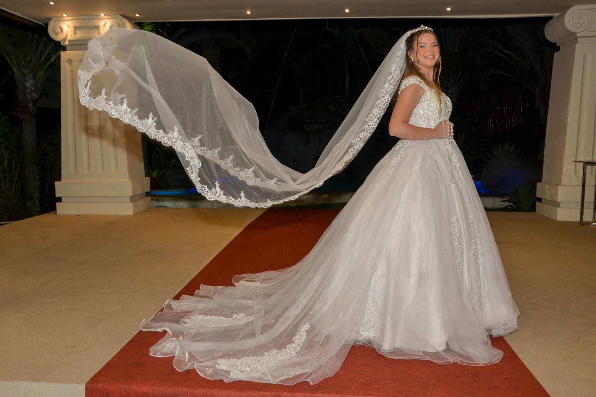 Noiva de vestido branco, véu longo esvoaçando, em pé no tapete vermelho.