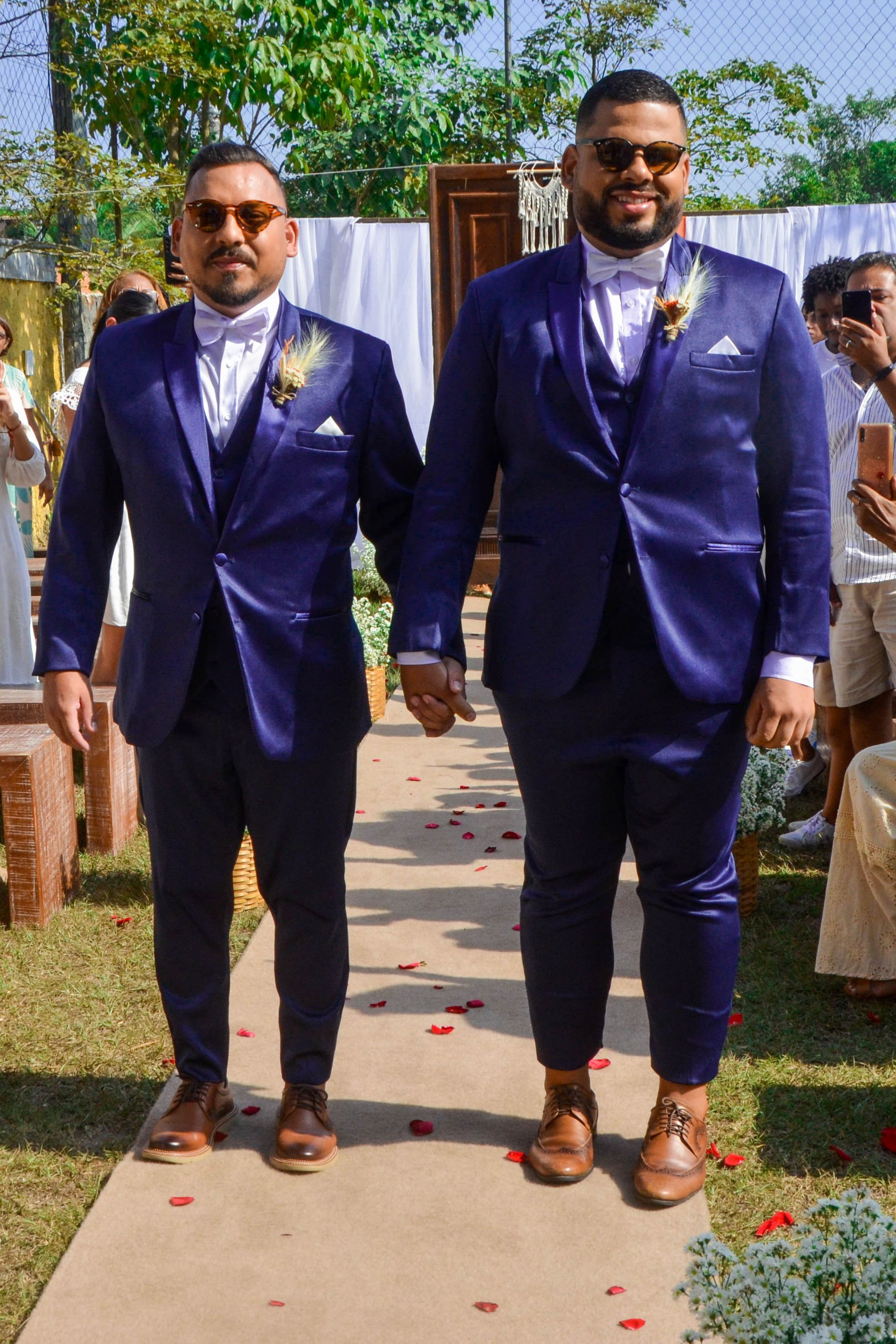 Dois homens de terno azul de mãos dadas, caminhando pelo corredor, com arranjos florais visíveis.