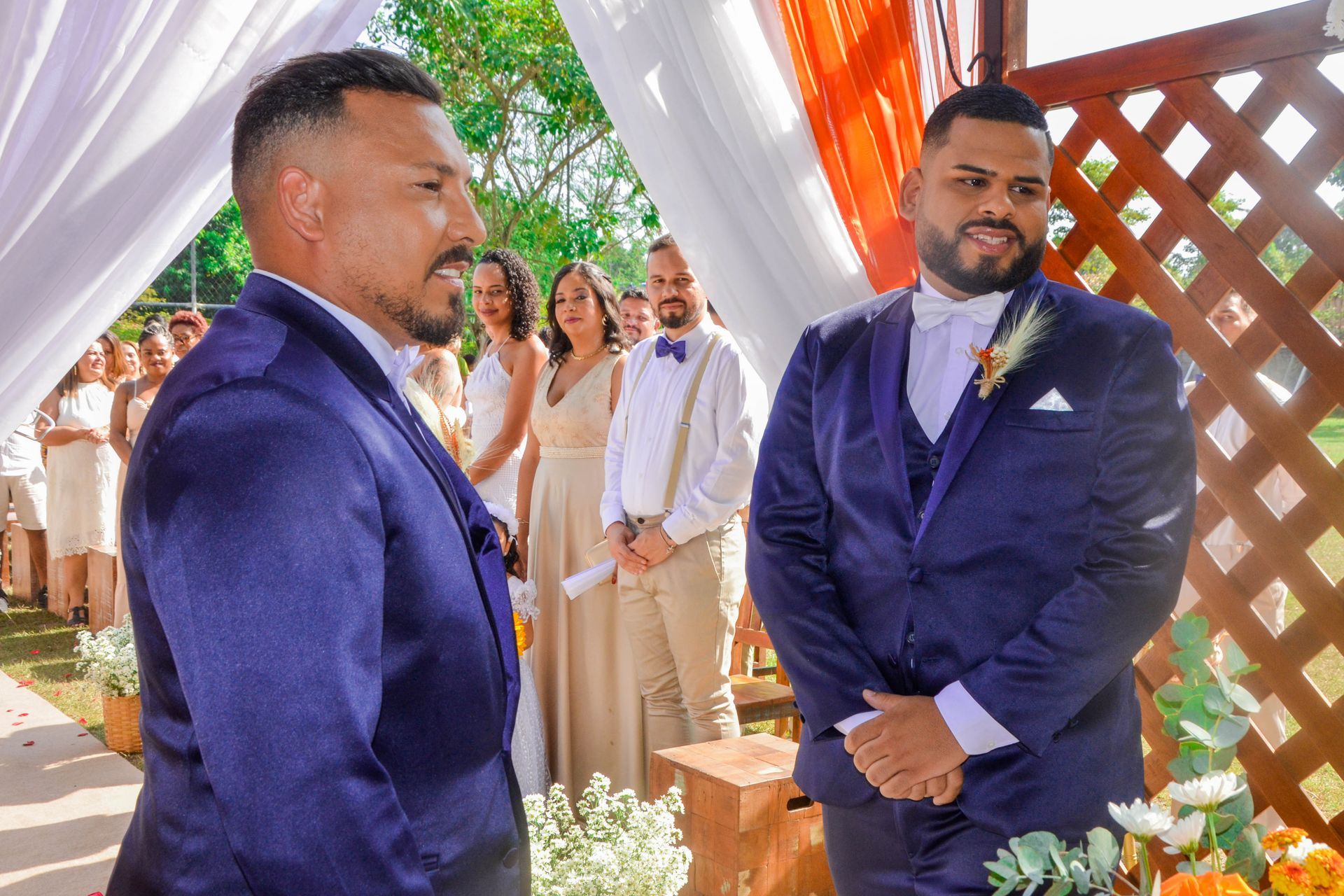 Dois homens de terno azul em uma cerimônia de casamento ao ar livre, um de frente para o outro, com a festa de casamento ao fundo.